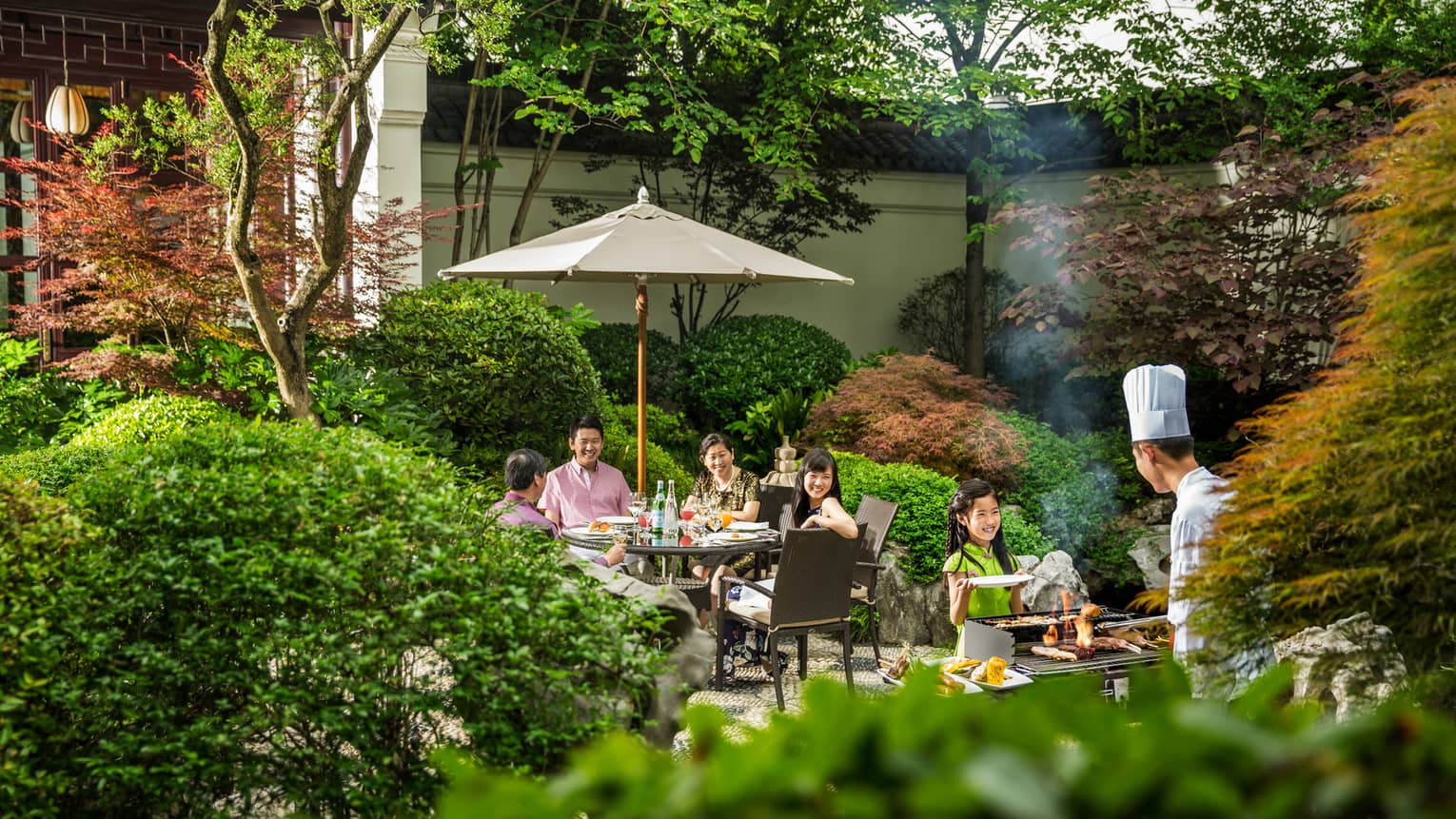 View over hedges, trees to family dining on patio, chef at barbecue