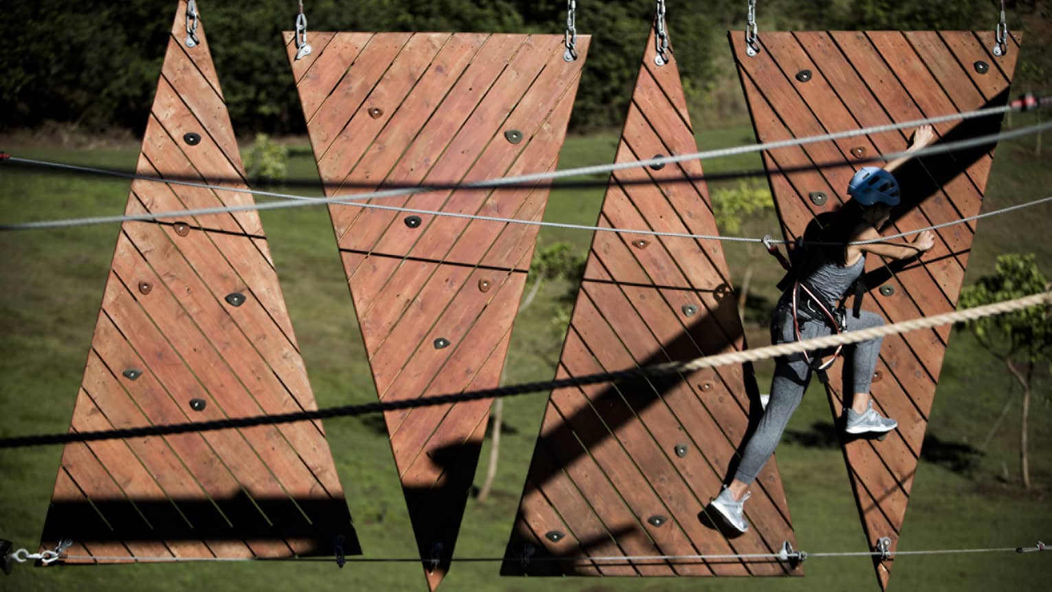 Rear view of person using climbing holds to traverse from one suspended climbing obstacle to another, green hillside beyond.