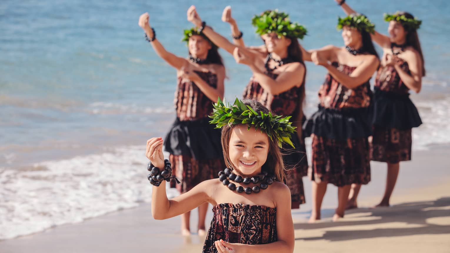 Hula dancing, smiling young girl, group of women behind her on beach, wearing traditional floral crowns, dresses