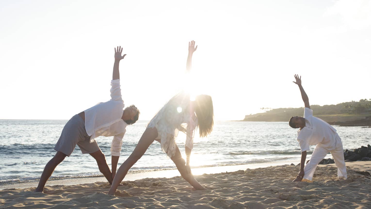 Two people in white beach shirts and shorts do a yoga pose with yogi at sunrise on beach