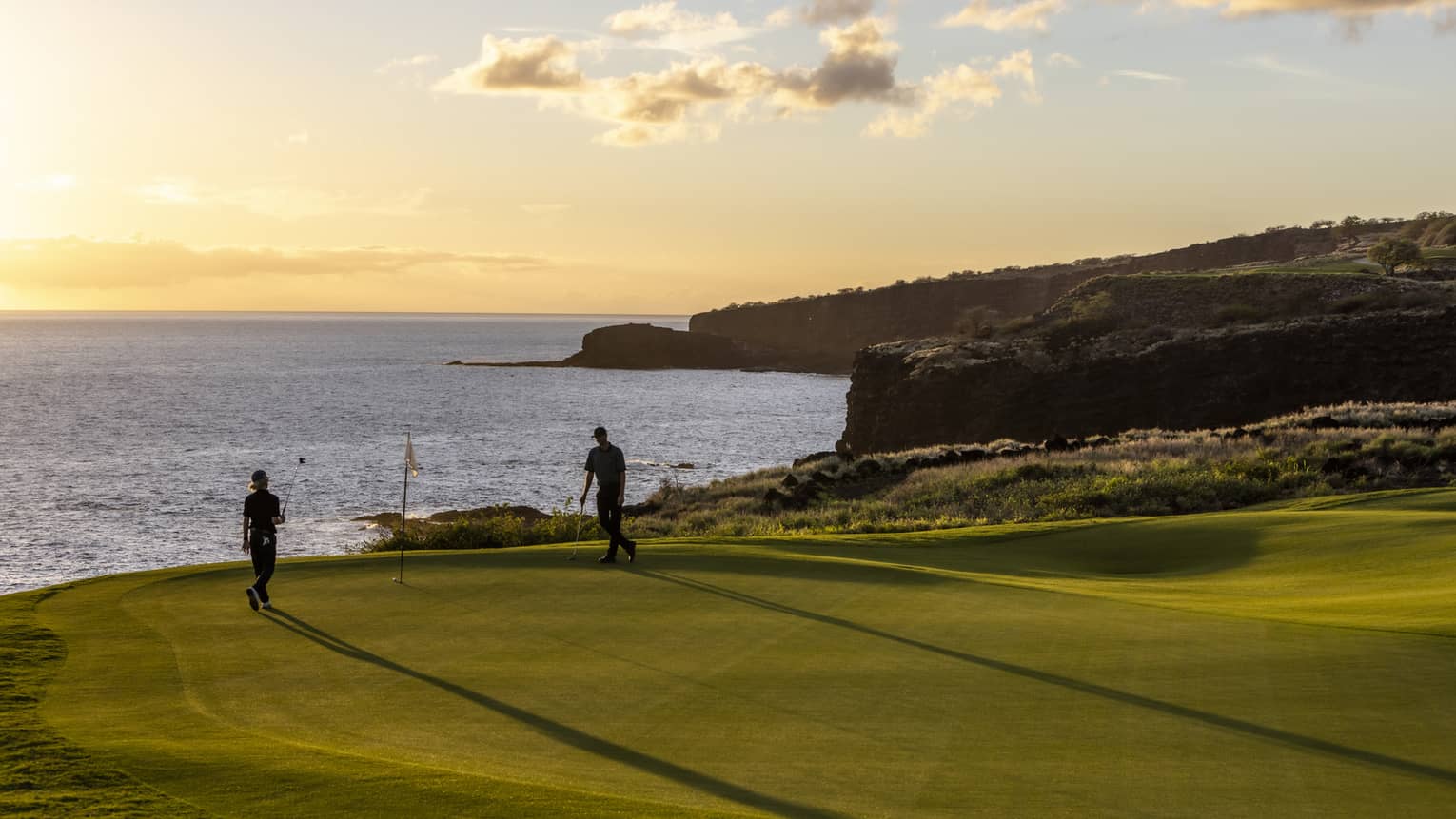 The setting sun silhouettes a pair of golfers enjoying a game of golf. The golf course is on top of a cliff beside the ocean.