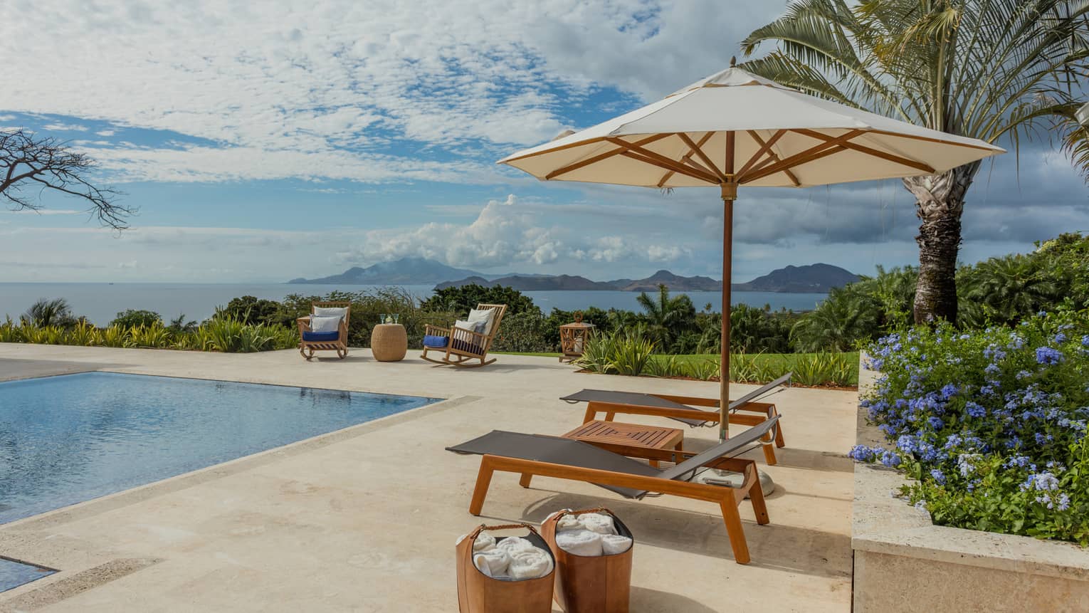 Two lounge chairs under patio umbrella, baskets with towels by outdoor pool