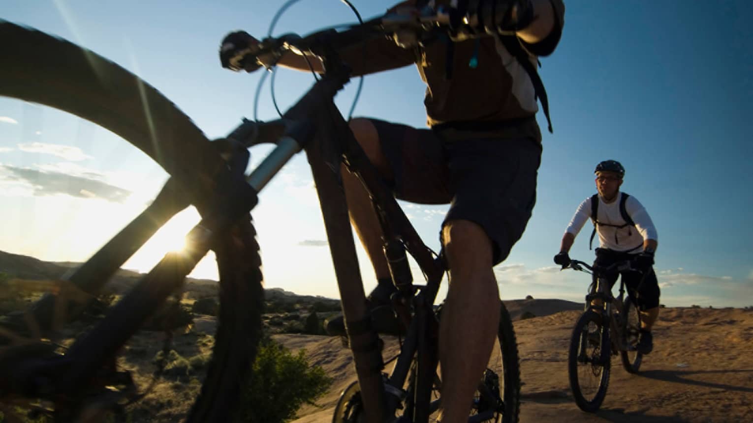 Close-up of cyclist in gear and helmet looking up from bike, second cyclist nearby as sun shines from behind