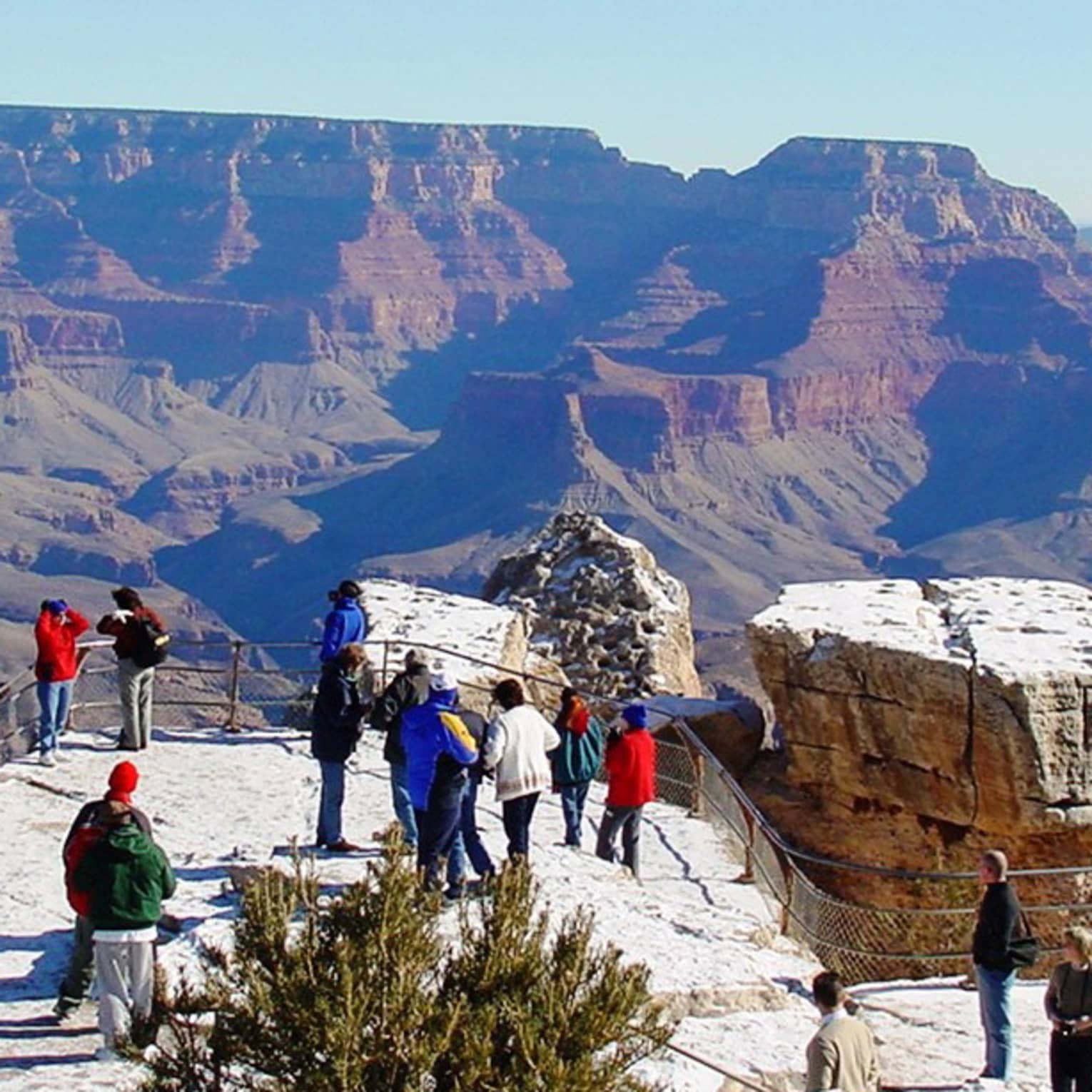 Tourists on a fenced platform, some taking photos, dwarfed by the the majestic Grand Canyon backdrop beneath a clear sky.