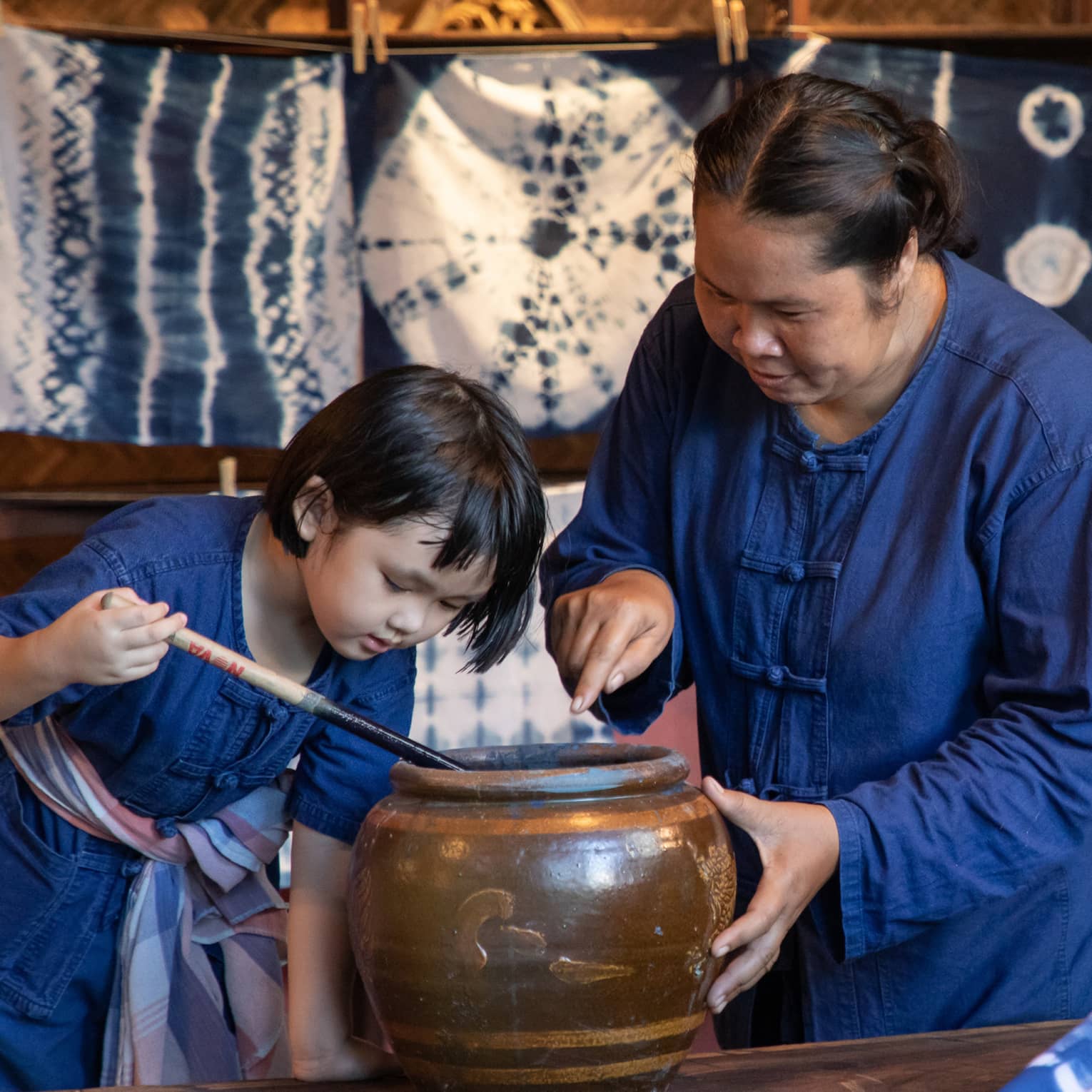 A woman teaching a child how to dye fabric using indigo, with tie-dyed cloths hanging in the background in a traditional workshop.