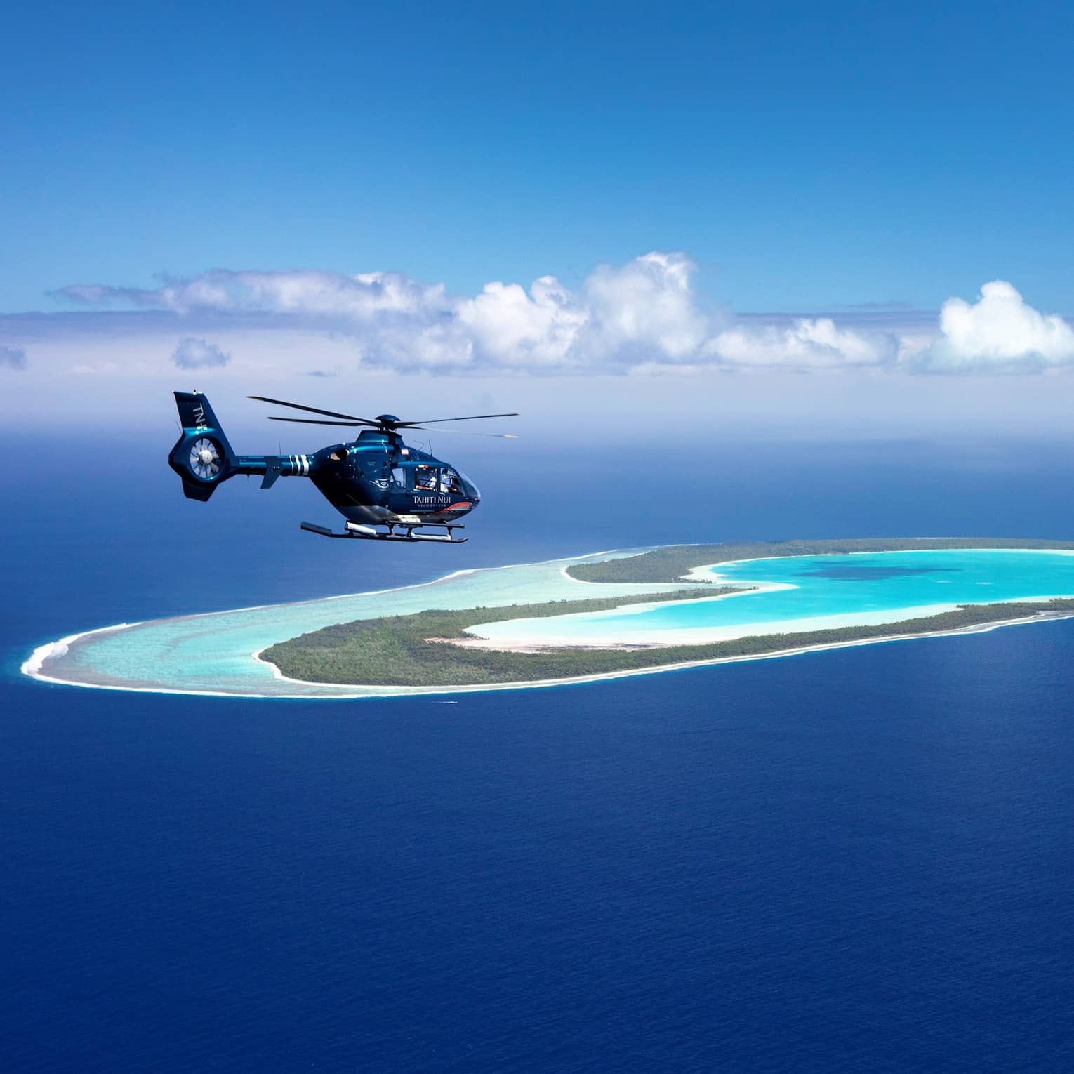 A helicopter flies over a teal lagoon, a heart-shaped island in the distance surrounded by endless ocean and sky.