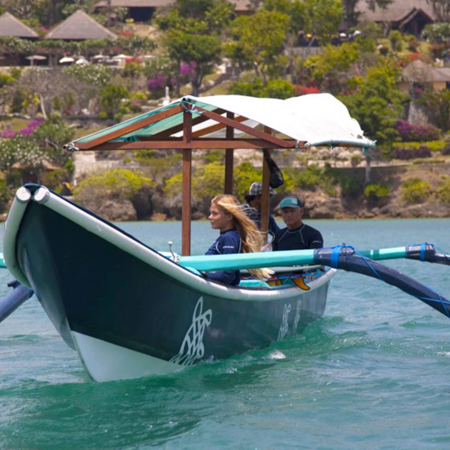 A guide sits at the stern of a covered outrigger canoe as two guests gaze at the water; behind, trees and huts dot the shore.