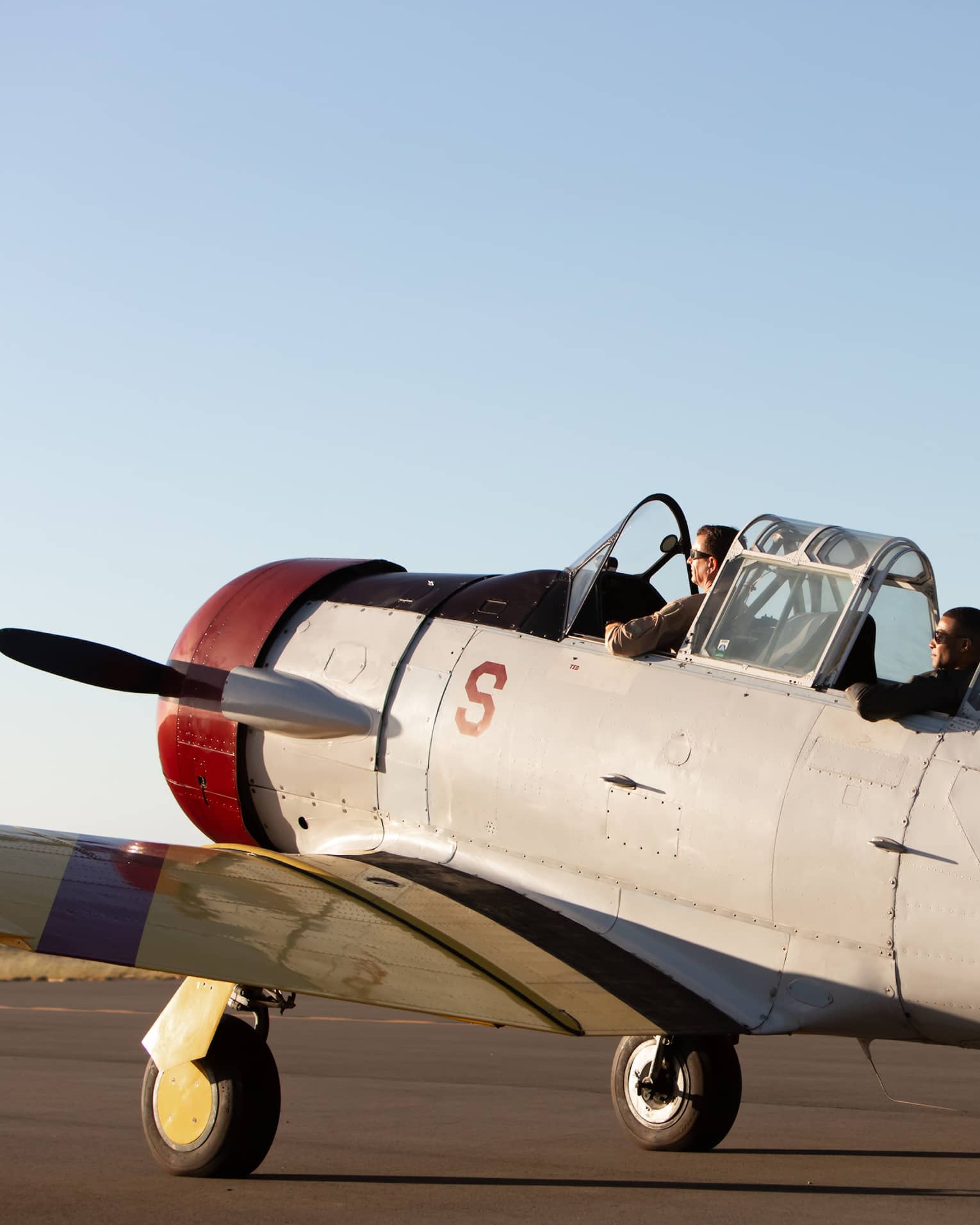 Two people sit in the cockpit of a vintage U.S. Navy aircraft on a runway, preparing for takeoff under a clear sky.