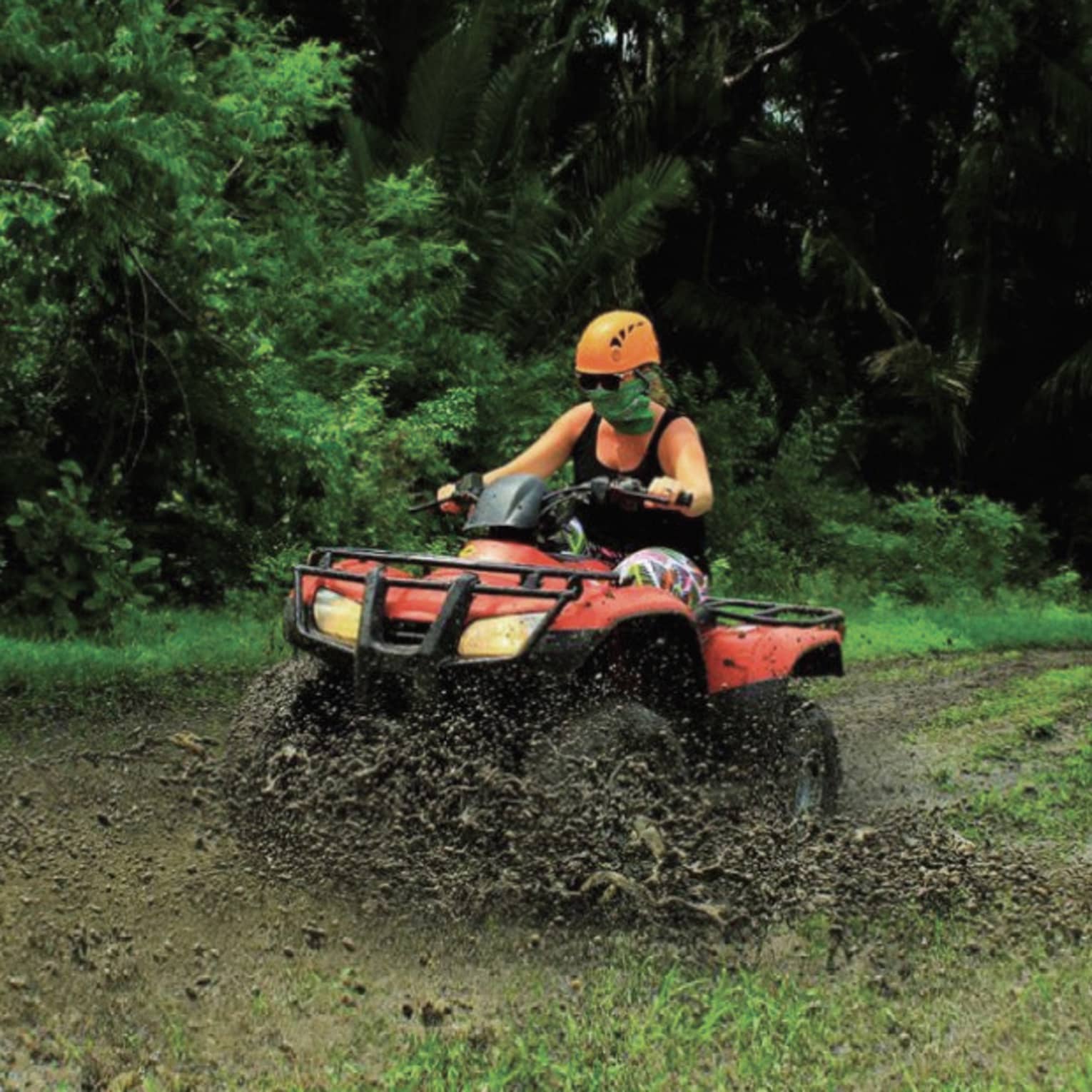 Person wearing a black tank top and bright orange helmet rides through the mud on a red ATV