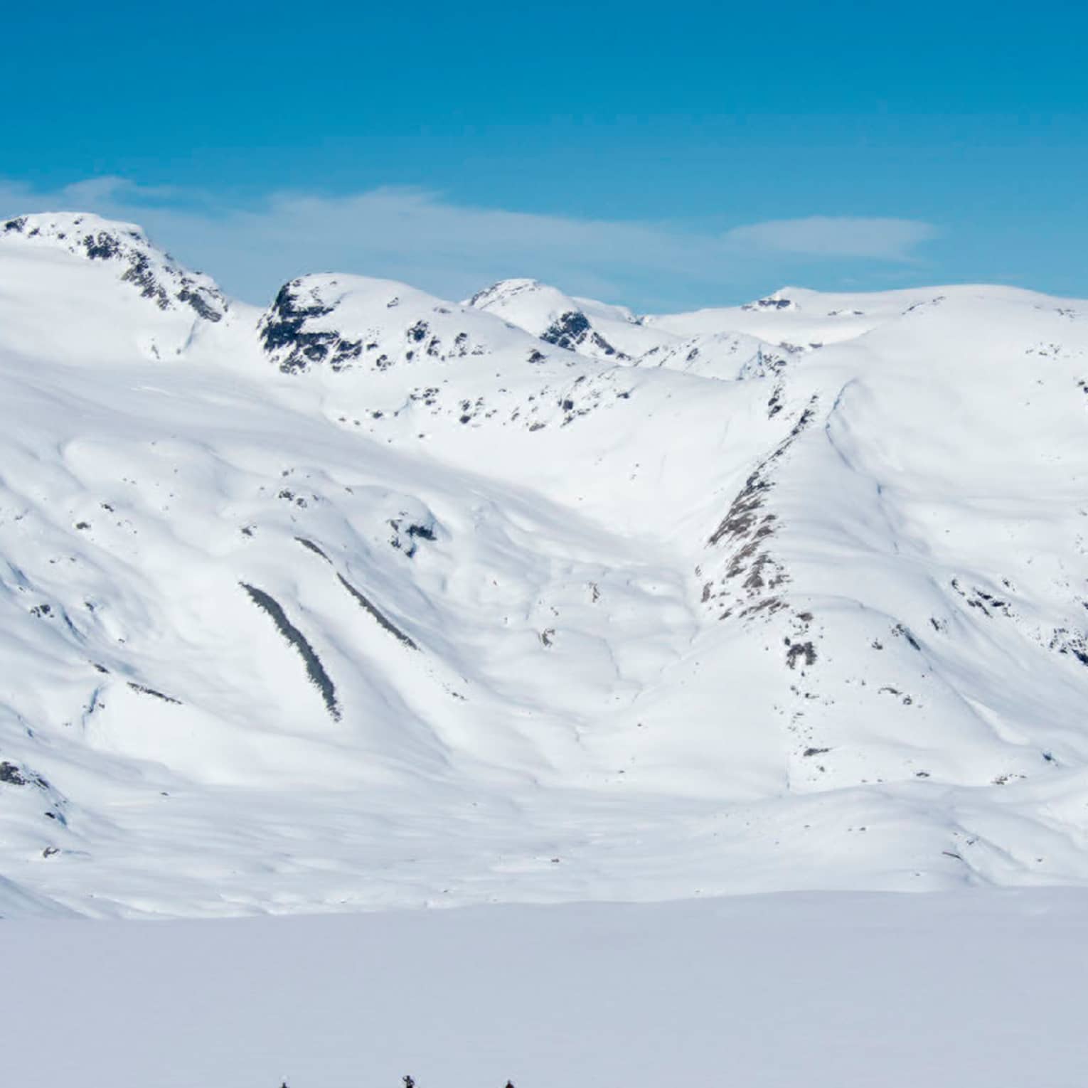 Tiny silhouettes of three skiers near top of snowy rocky mountain, blue sky