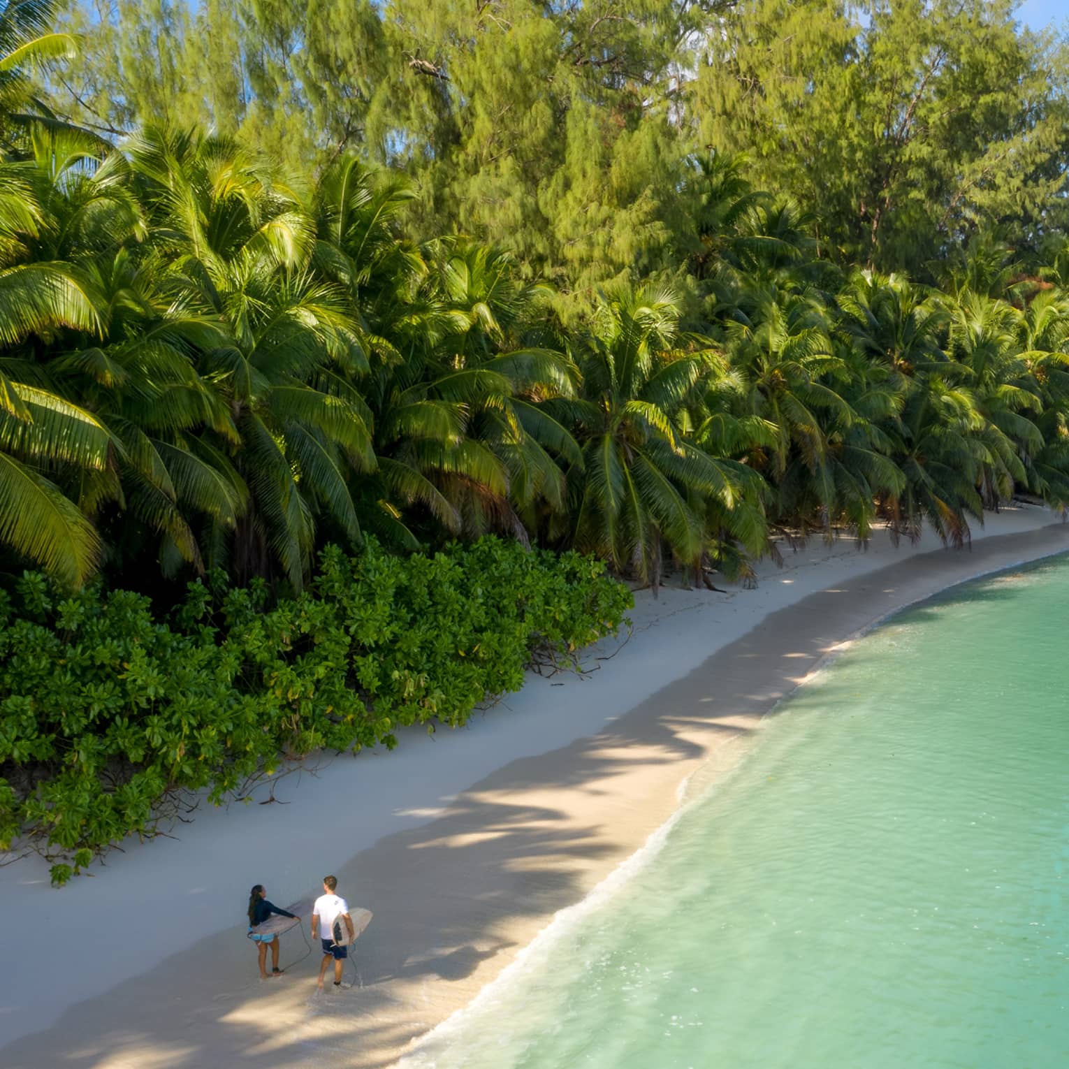 Couple walking along beach, carrying surfboards, flanked by lush tropical vegetation and blue-green water