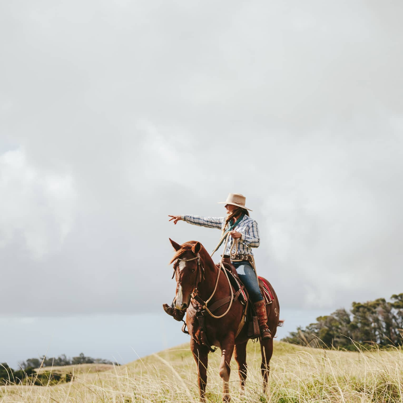 A couple riding horses, looking out at the long grass fields, with grassy hills and trees in the distance on a cloudy day.