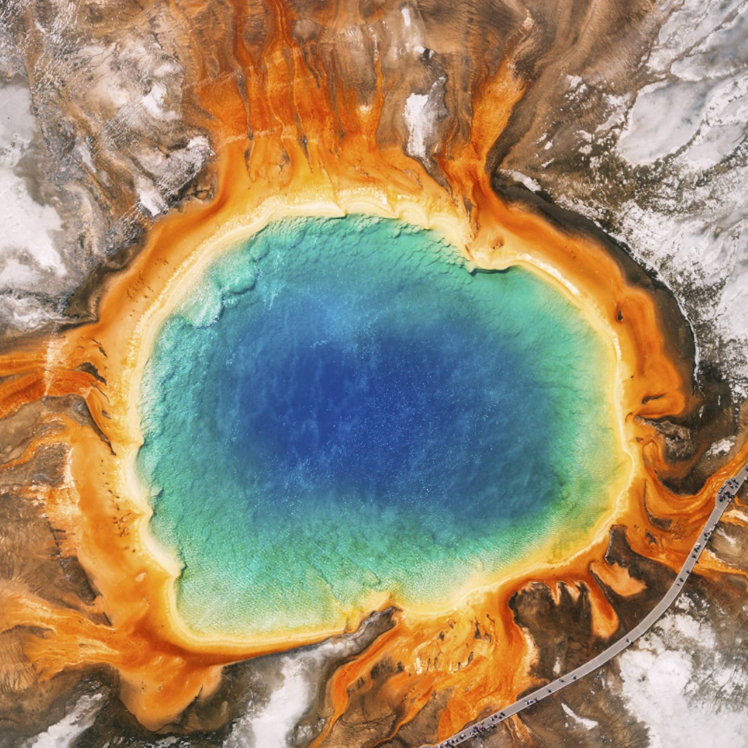 Aerial view of the Grand Prismatic Spring in Yellowstone, featuring vibrant blue, green, yellow and orange concentric rings, surrounded by rocky terrain