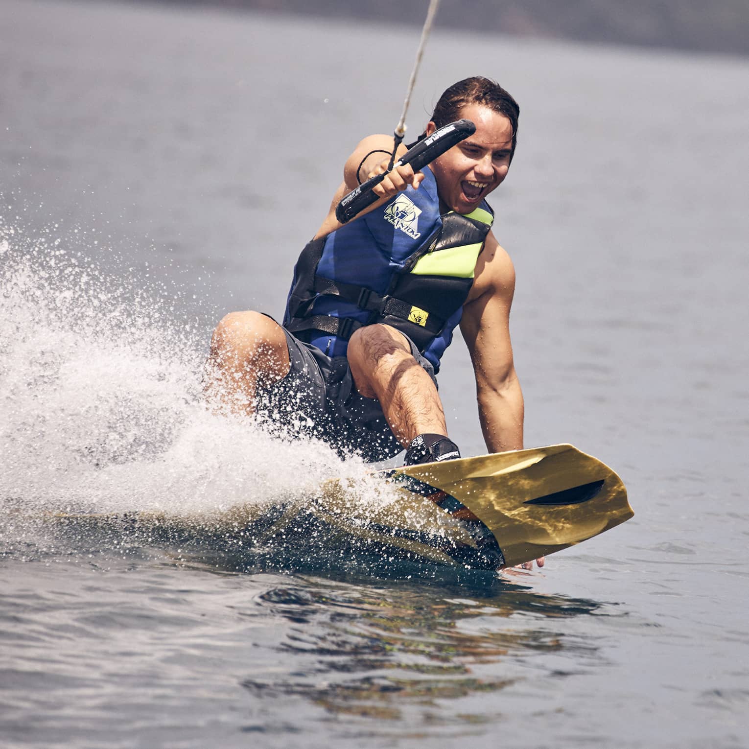Young man wearing a blue and green life vest and blue swim trunks wakeboarding through the water