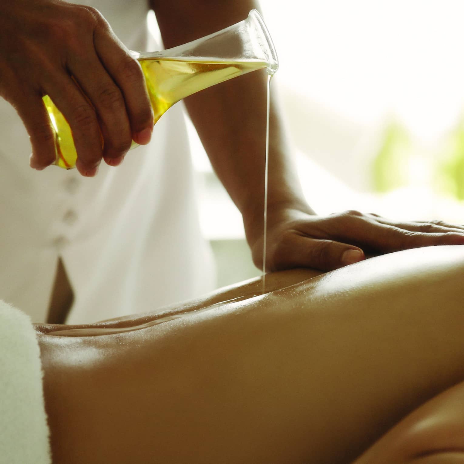 Spa staff pours massage oil over woman's bare back as she lays on treatment bed