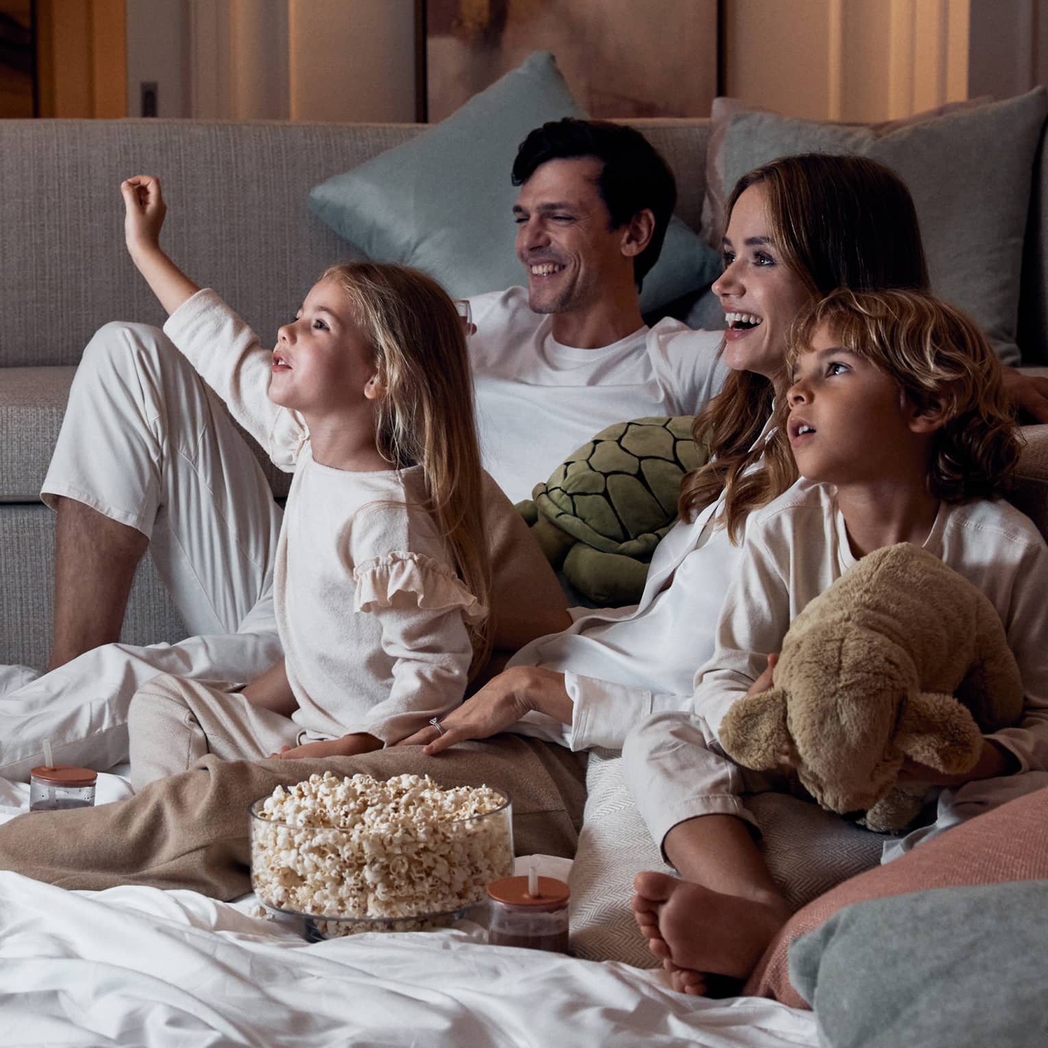 A family relaxing on blankets beside a sofa; the children hold stuffed toys and there's a bowl of popcorn between them.
