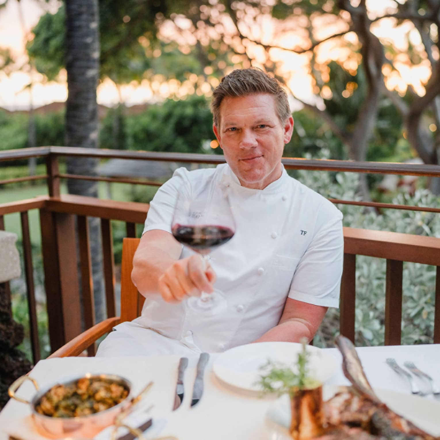 A smiling Tyler Florence sitting out on a veranda wearing his chef jacket and giving a toast with a glass of red wine.