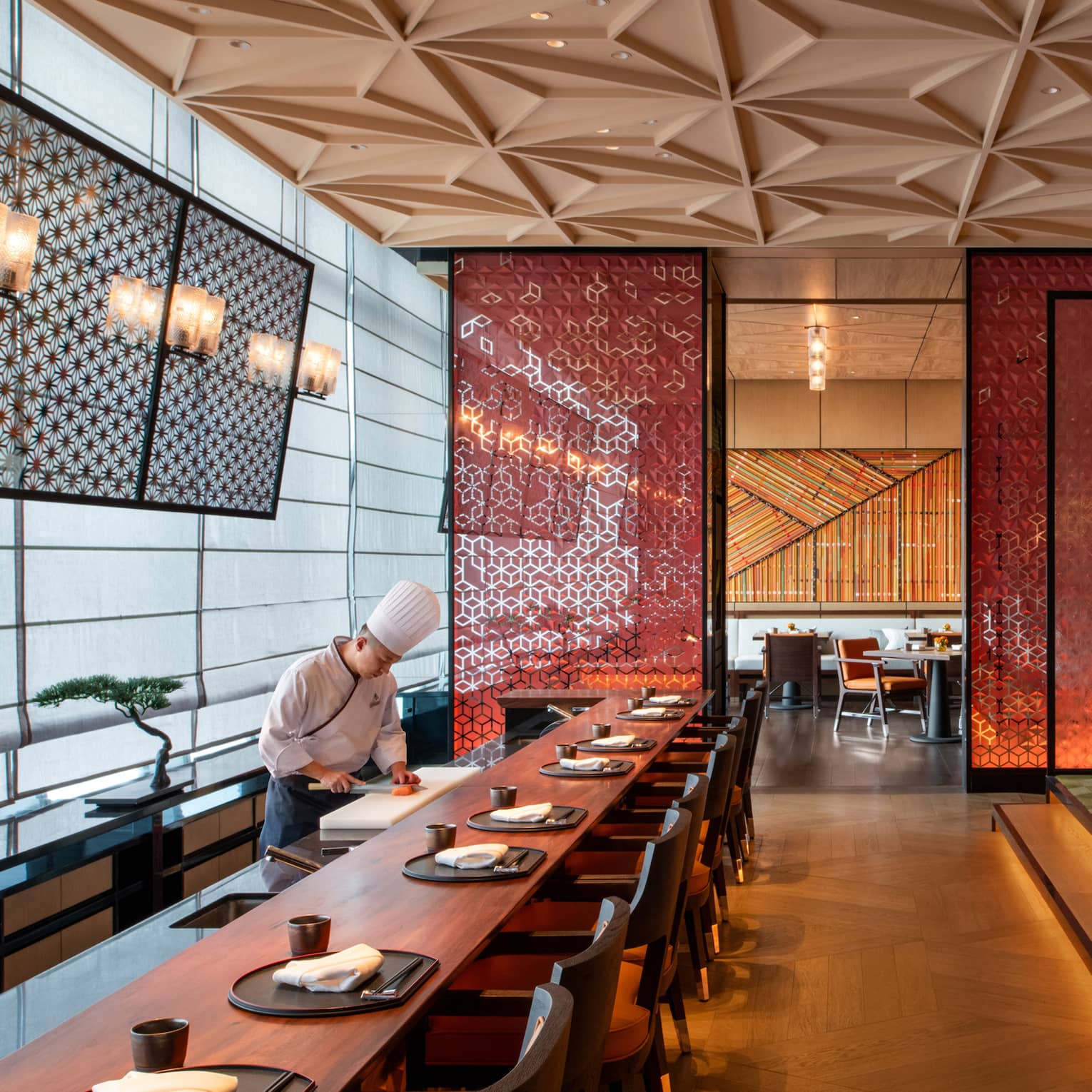 A sushi chef works by a long wood dining table in a modern restaurant, a red ornate screen and walls reflecting golden light.