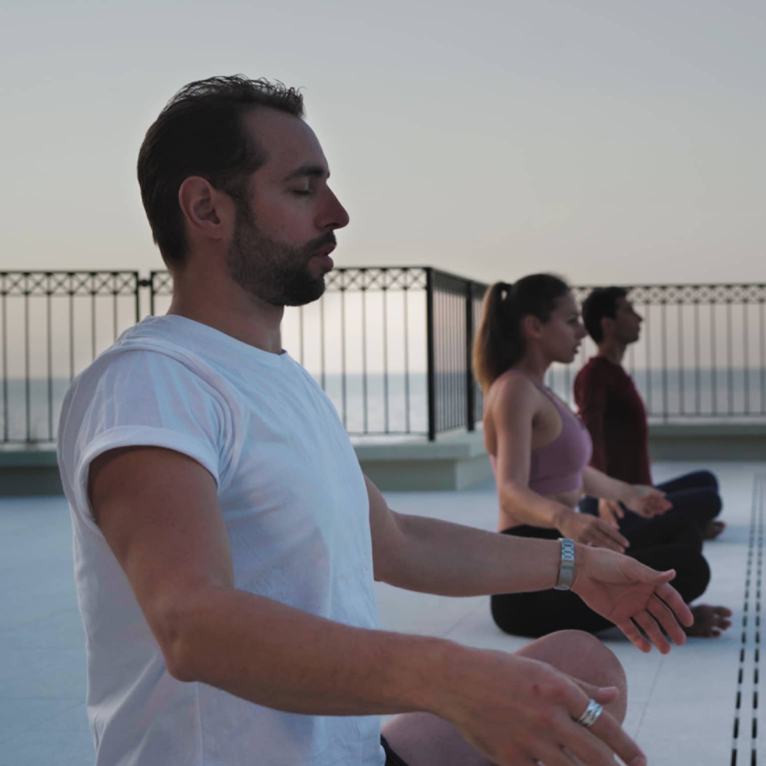 Under a grey sky, three guests sit cross-legged with eyes closed beside a calm pool bordered by greenery and the vast ocean.