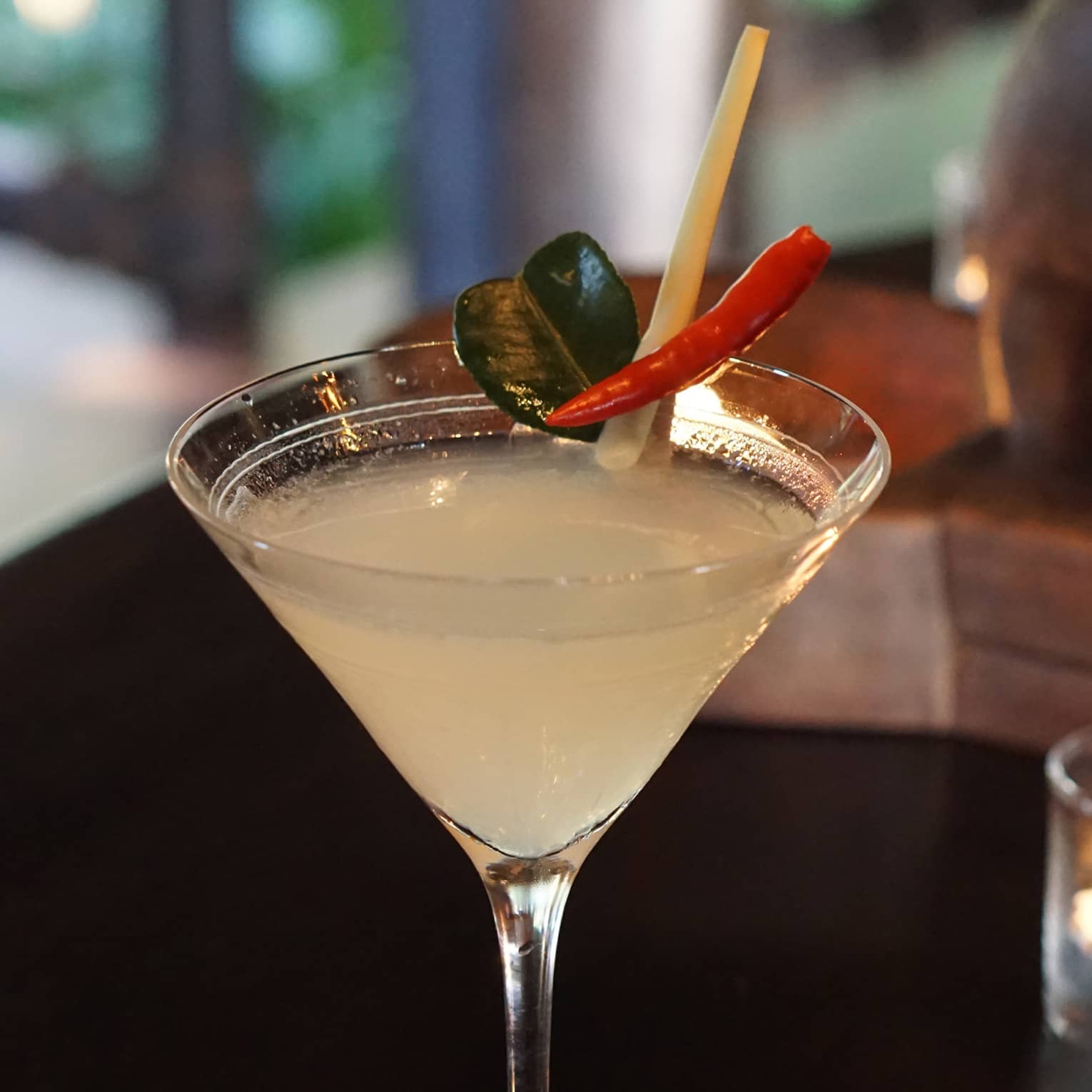 Martini glass with a citrus cocktail garnished with chilli, lemongrass stalk and makrut lime leaf