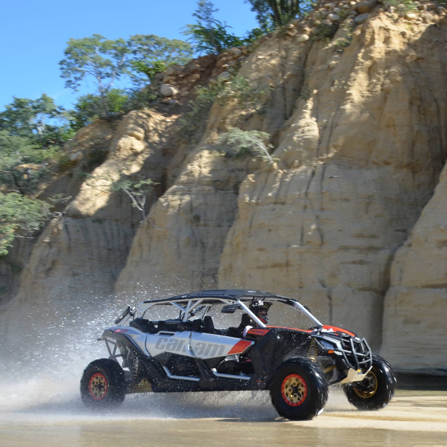 Water sprays off the back of a sporty off-road vehicle racing past deep vertical fissures in a tall crag topped with trees.