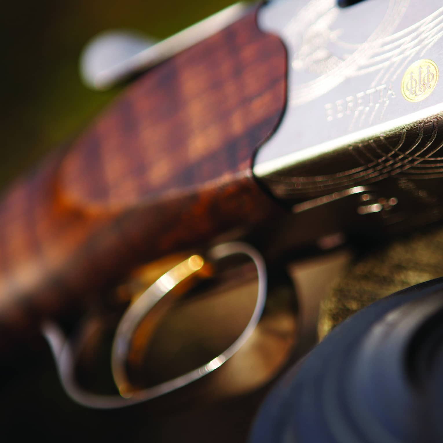 Close-up of trigger and wooden barrel of a rifle with silver engraving of a grouse, the Beretta name, swirls and a gold logo.