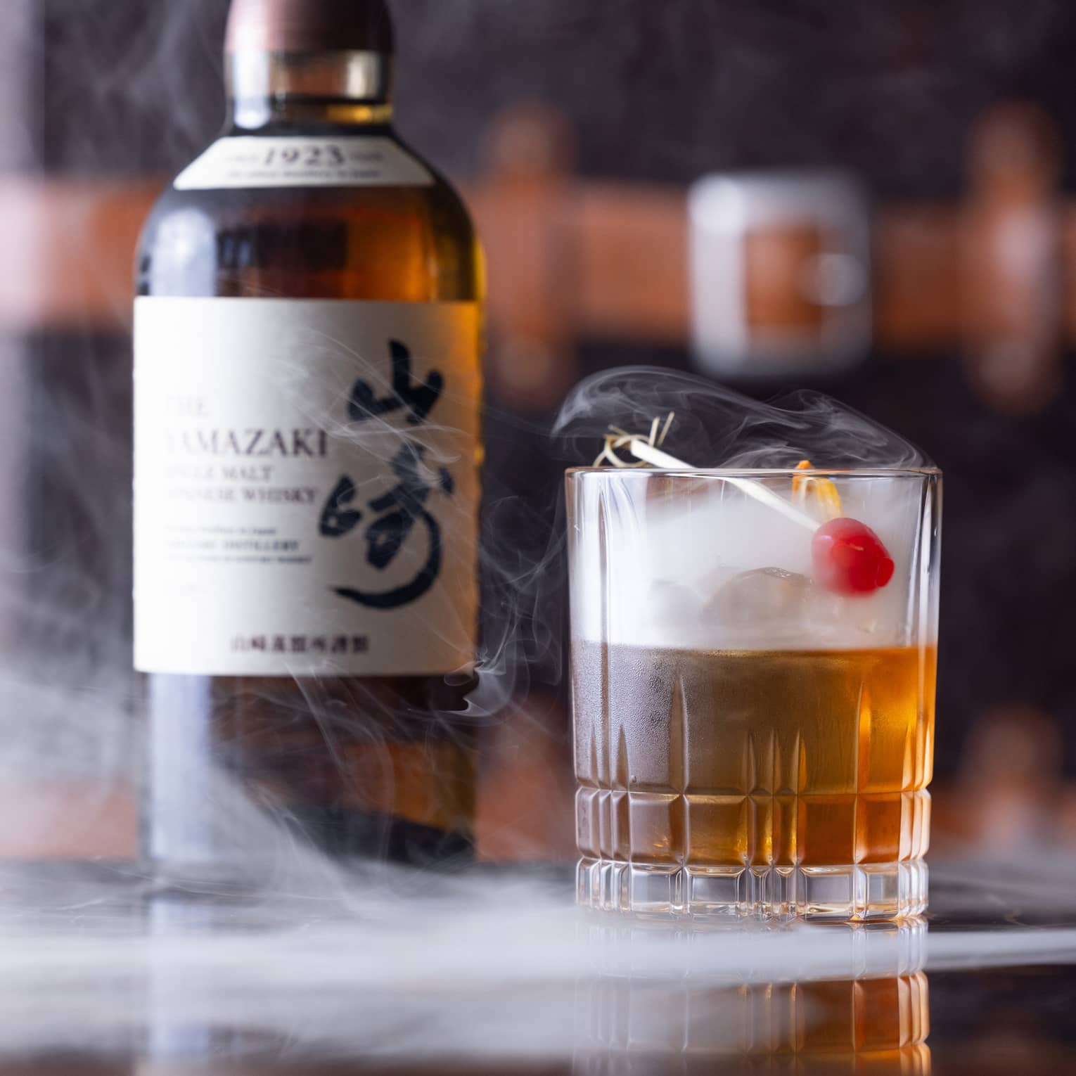 Bottle of Japanese whisky beside half-full cut-crystal glass with red cherry garnish, dry ice mist swirling from the glass.