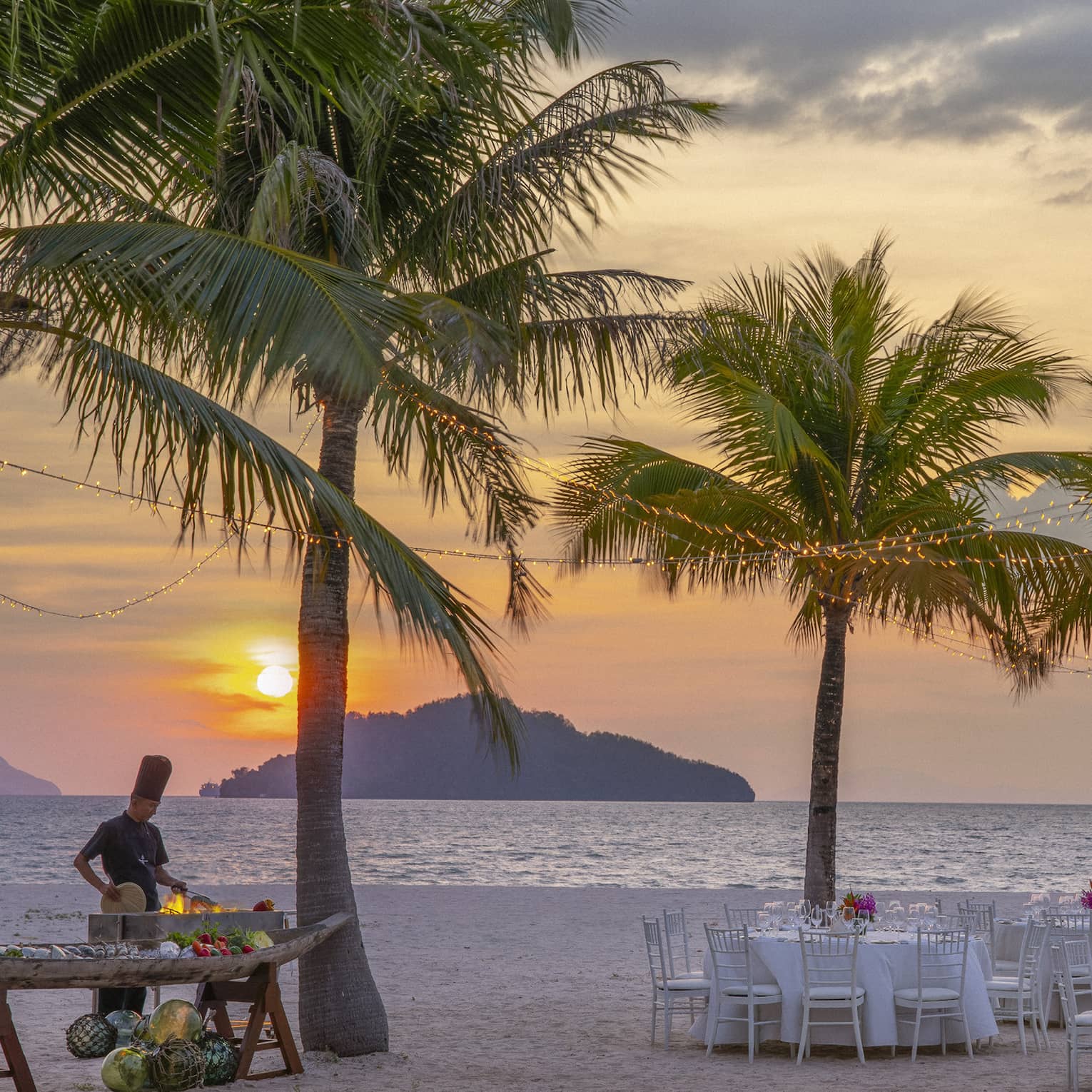 Tables set for dinner on the beach under a canopy of palm trees, a chef cooking over an open flame against the setting sun.