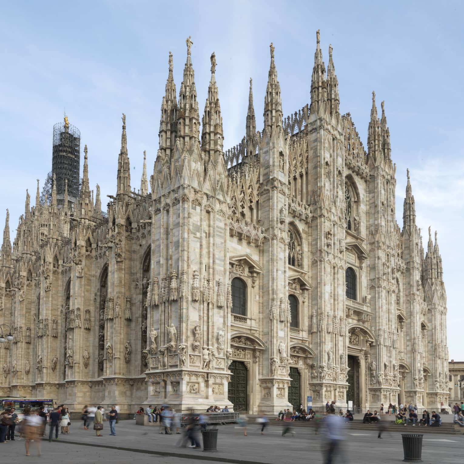 The exterior of the Milan Cathedral (Duomo di Milano), showcasing its intricate Gothic architecture with tall spires and detailed stone carvings against a clear sky.