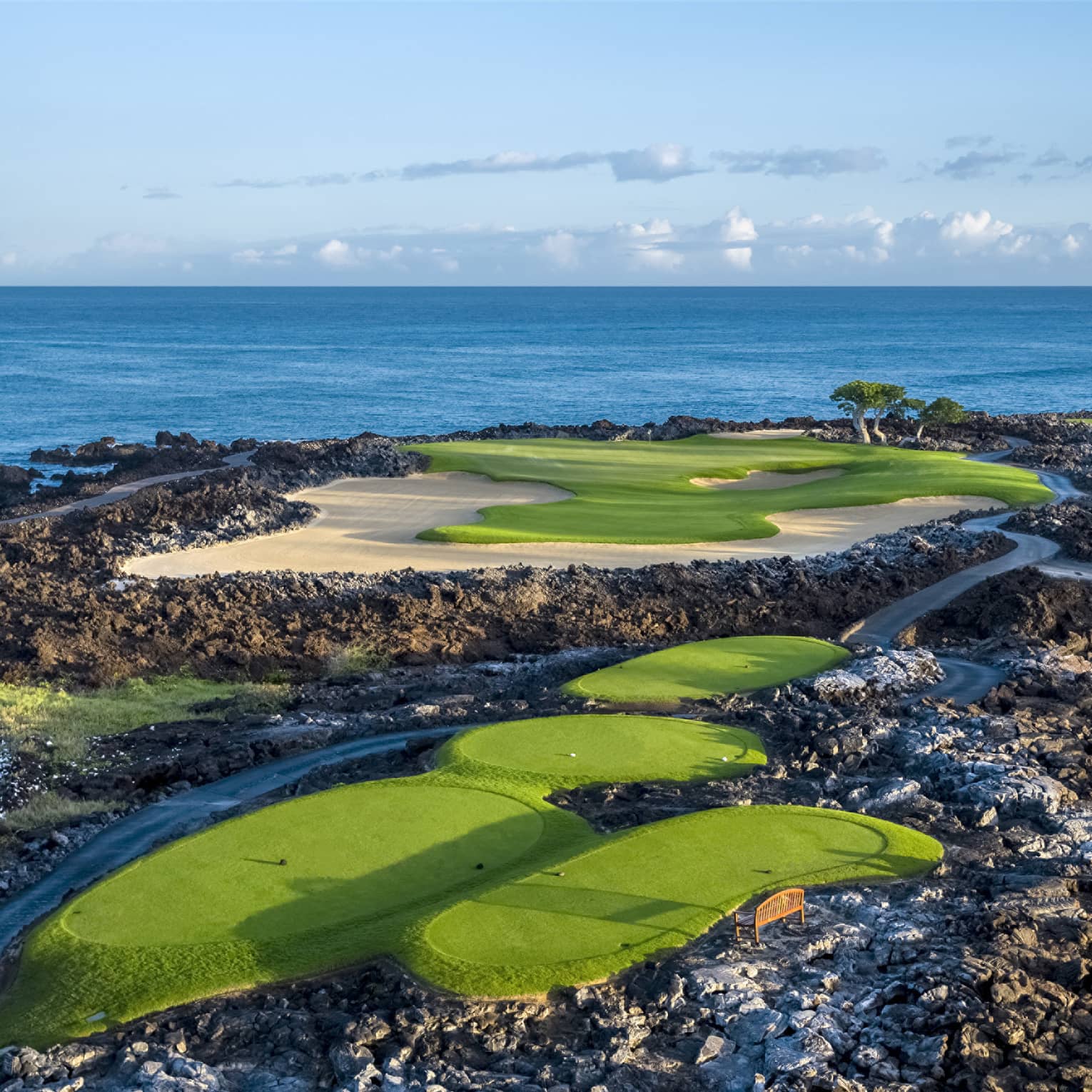 An impressive golf course overlooking the ocean, fairways and sand traps amid black lava formations, blue skies overhead.