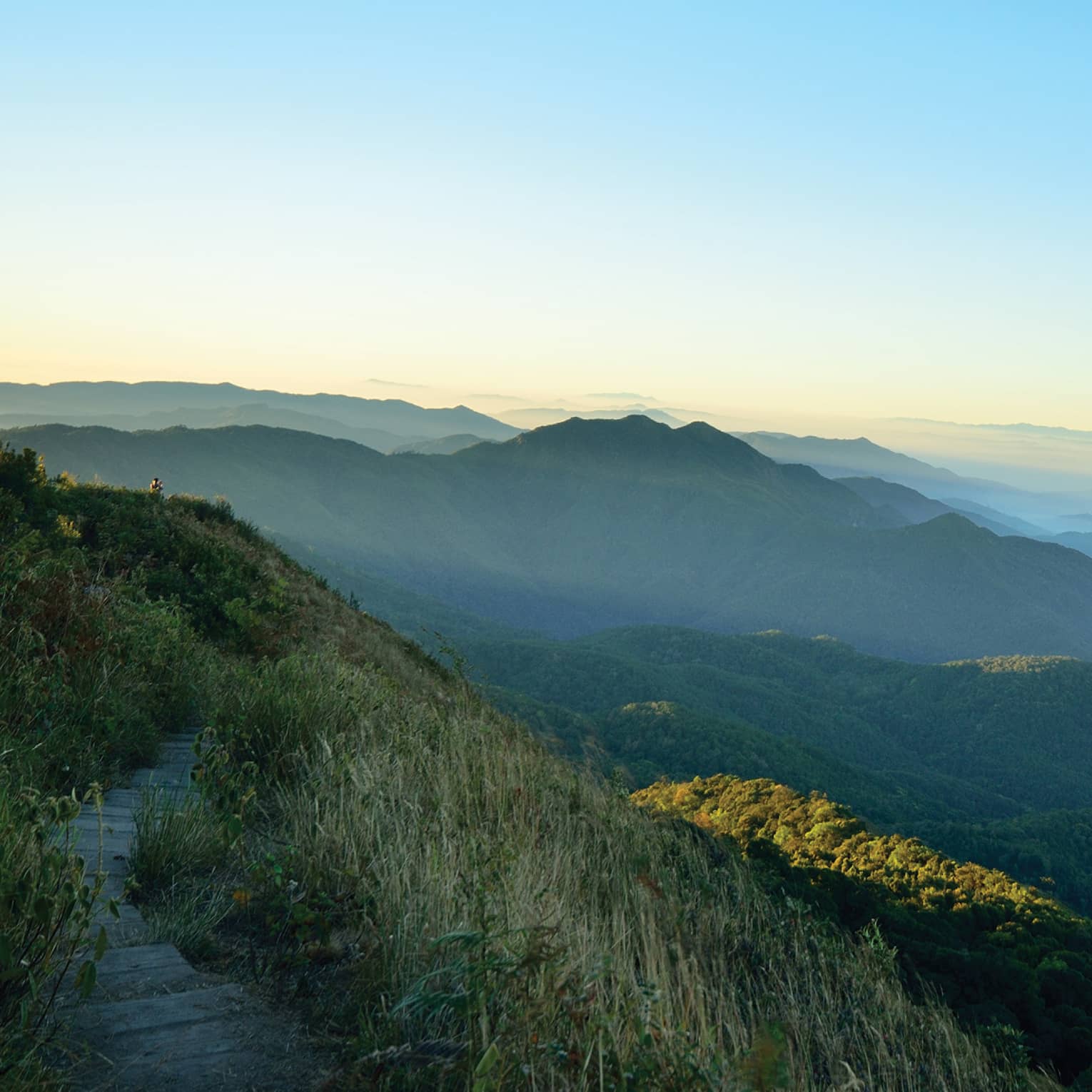 View from an elevated grass-lined path of low, forested mountains receding into the misty distance under blue and orange sky.