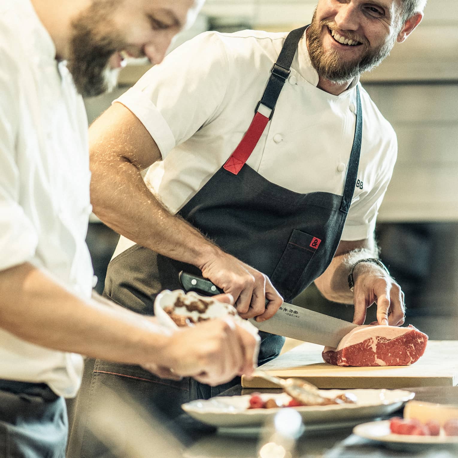 Two smiling chefs stand at a metal counter, one chopping a piece of meat while the other arranges food on a platter.