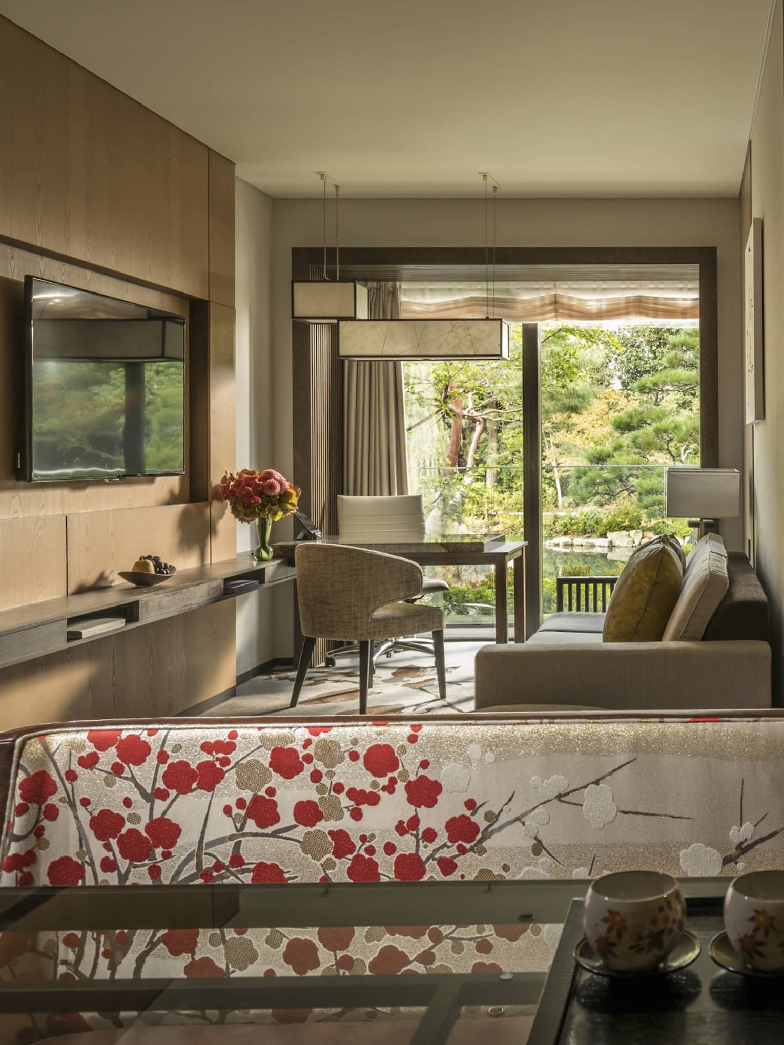 Connecting hotel room with sofa, desk and chairs, floral-patterned screen, tea set and large window overlooking a tranquil pond garden