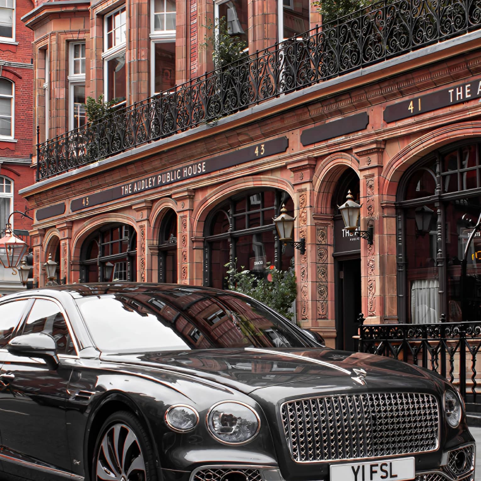 Close-up front view of shiny black modern Bentley with an elaborate grille parked in front of a restored Victorian building.
