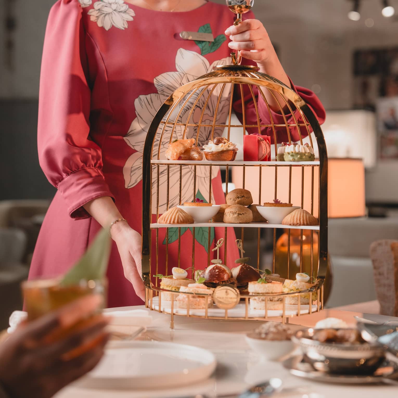 Server wearing a dark-pink long-sleeved dress places a three-tiered tray filled with sweet and savoury items for afternoon tea onto a table