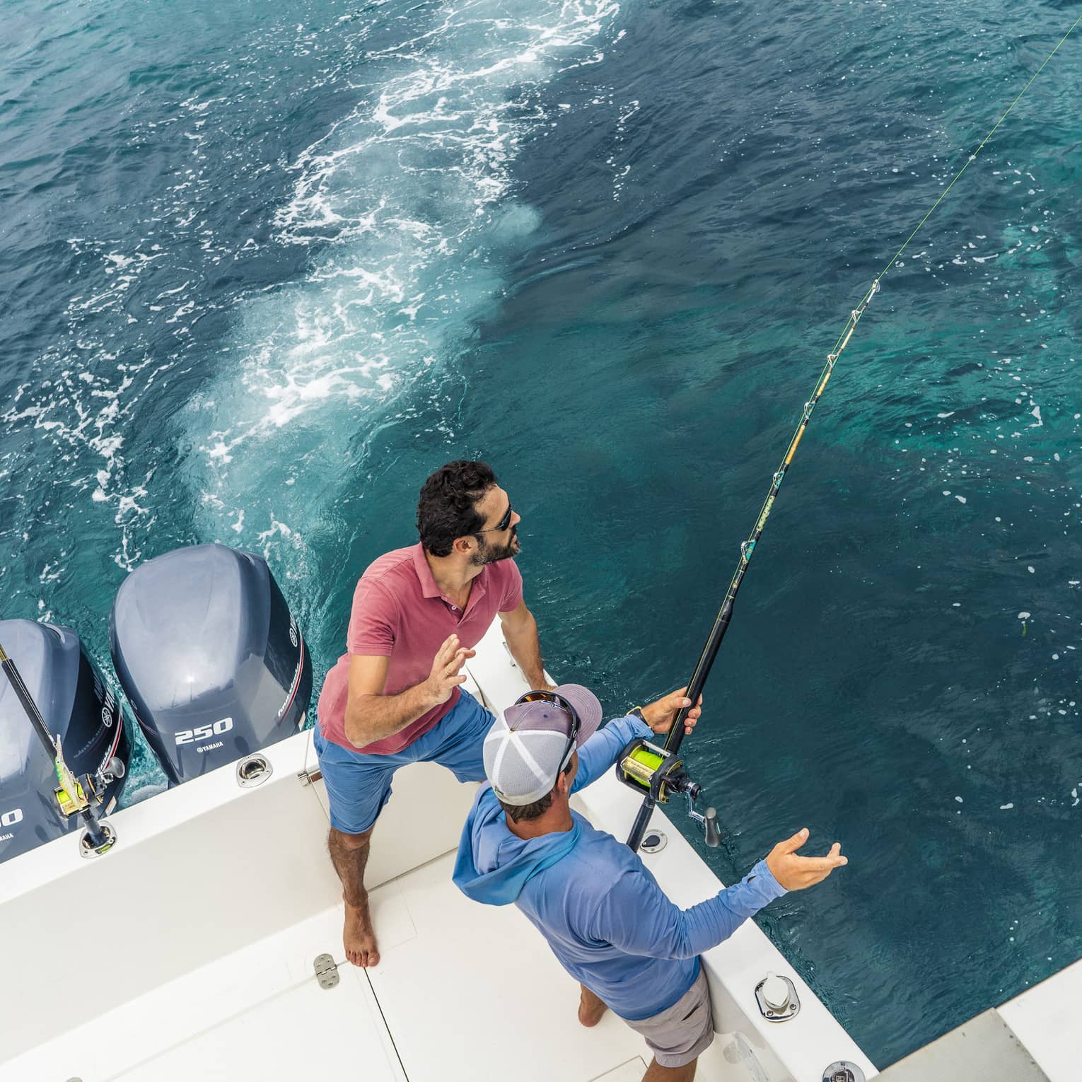 Aerial view of two people fishing off the back corner of a motorboat. One grips a fishing rod while the other looks on.