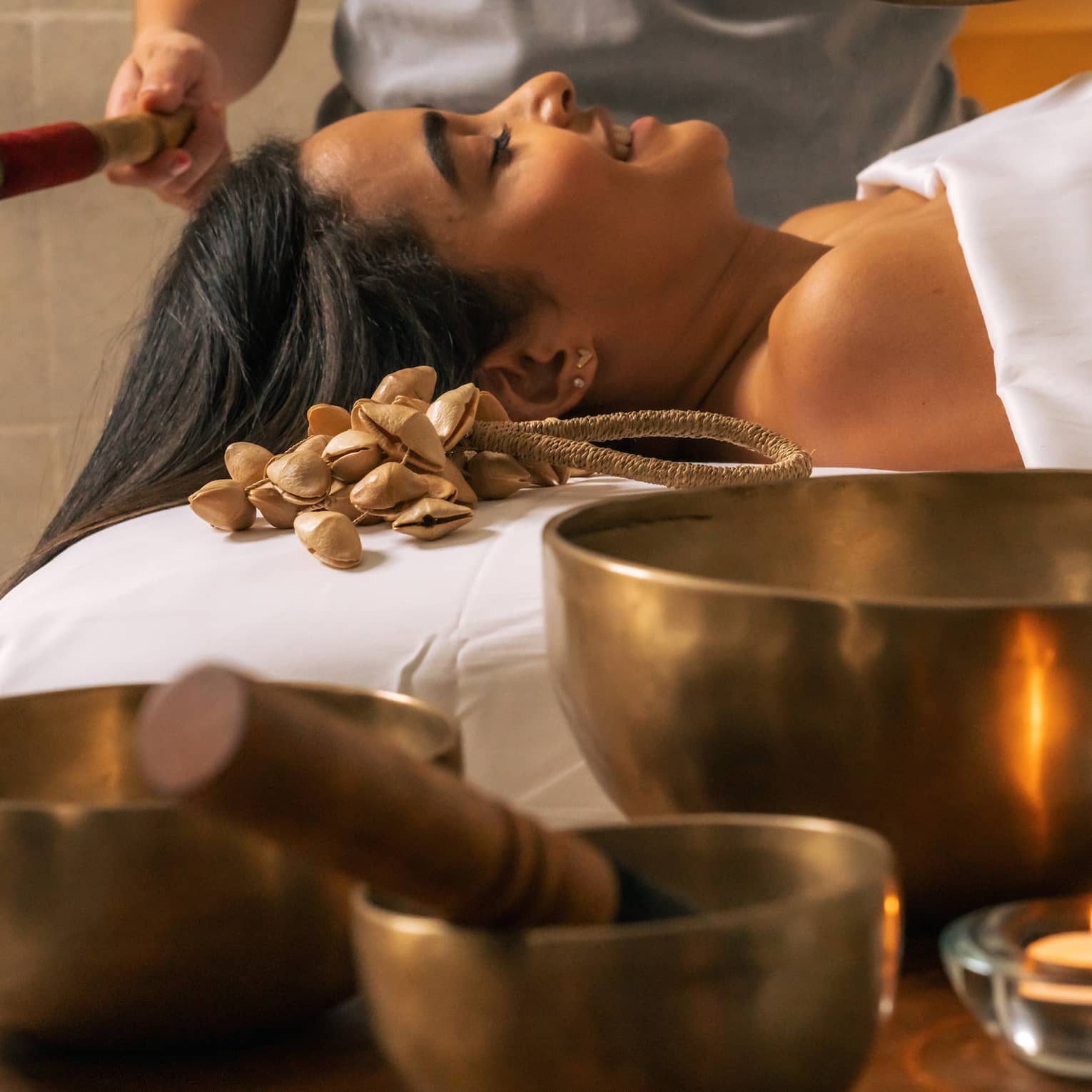 Three Tibetan sound bowls sit on a table next to a person lying beneath a white sheet while another person rings a smaller bowl over them