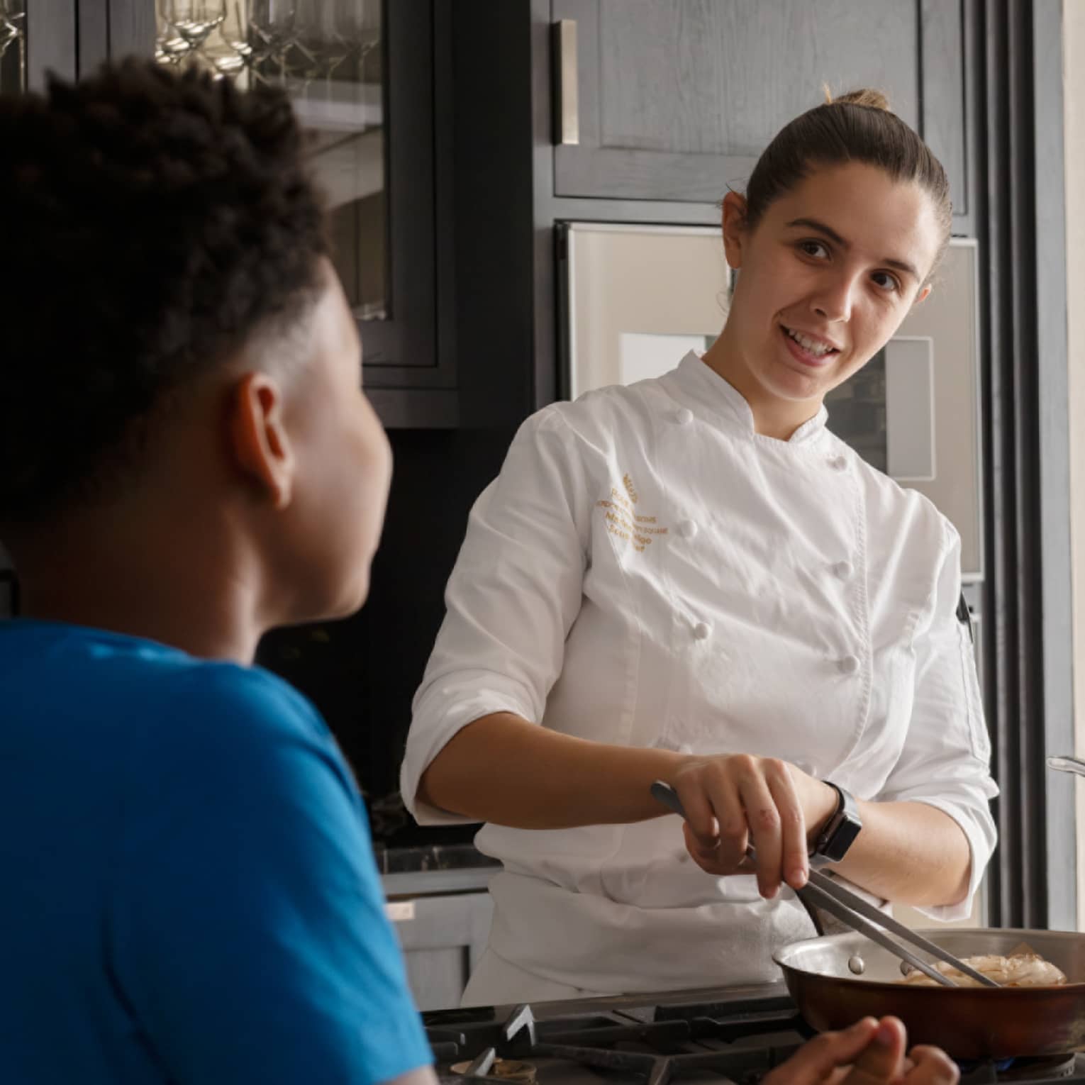 Rear close-up of a child looking on as a smiling chef prepares food in a copper frying pan, a copper pot on a nearby burner.