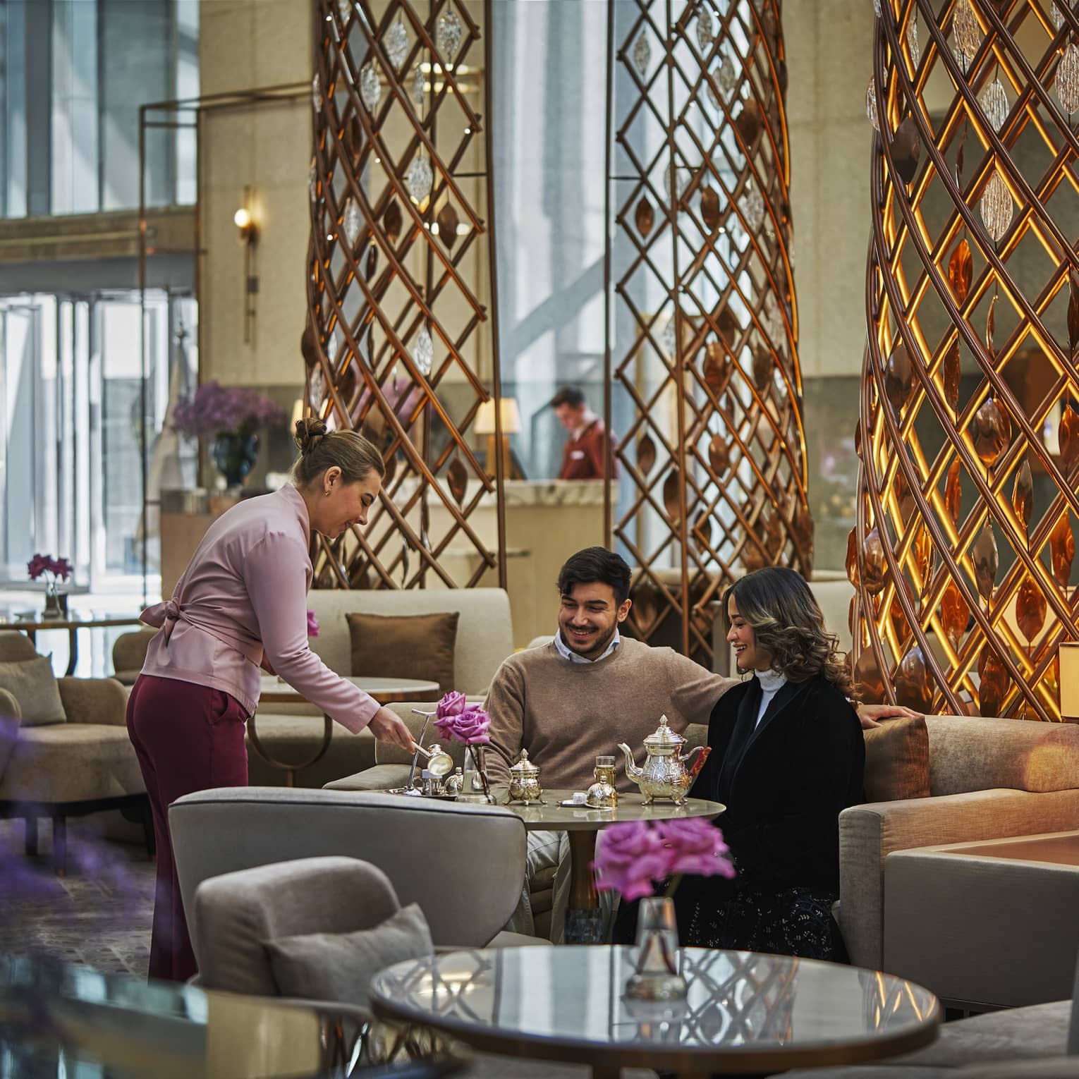 Two people sit a table in the middle of a lobby lounge while a server pours them tea