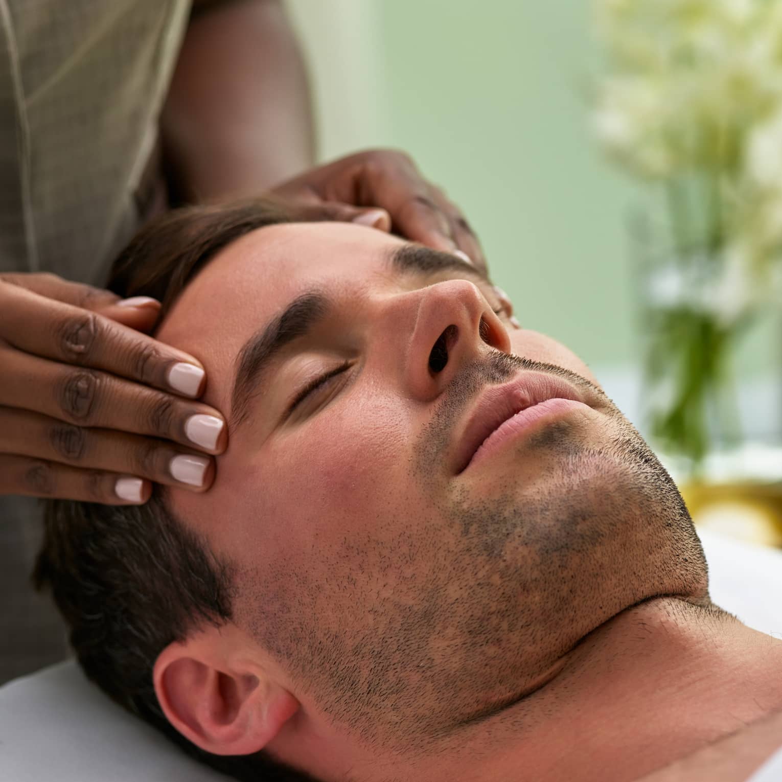Man lies on massage table with eyes closed as hands massage his temples, forehead