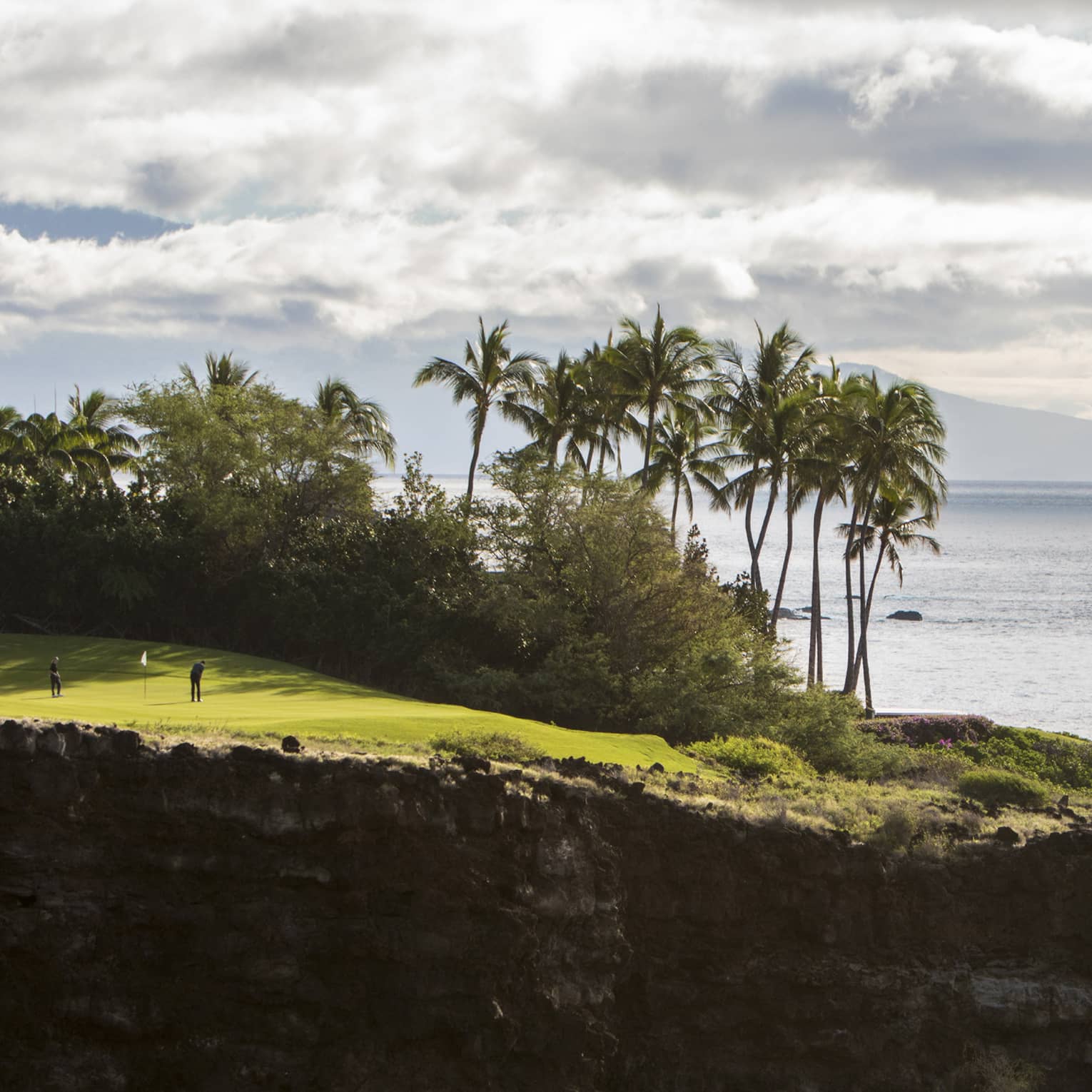 Long view of two people on clifftop golf green aside lush trees and towering palms; fluffy clouds, ocean and mountain beyond.
