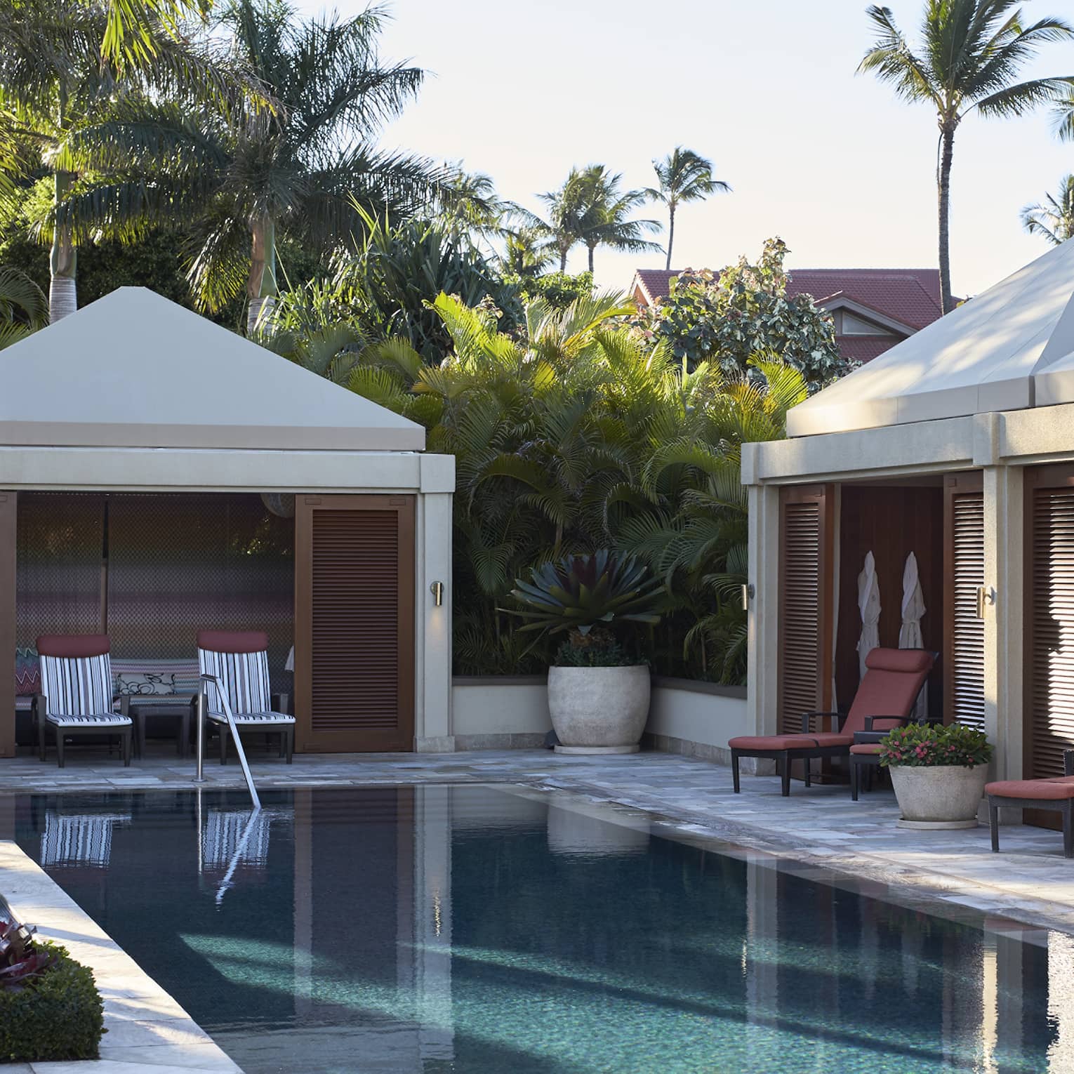 Cabanas line outdoor swimming pool deck under palm trees and sunny sky
