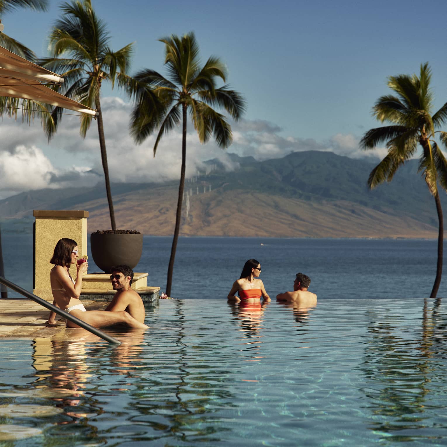 Four people relax in an infinity pool overlooking the ocean and palm trees with a mountain in the background