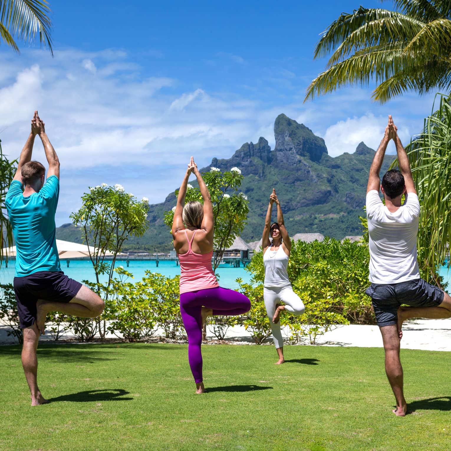 Three guests and an instructor practice yoga on the lawn, the lagoon and mountains visible behind them