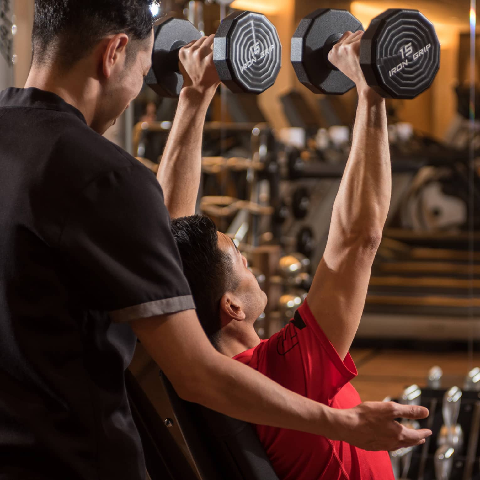 Man in chair in front of mirror lifts two 15 lb. weights above his head while personal trainer assists