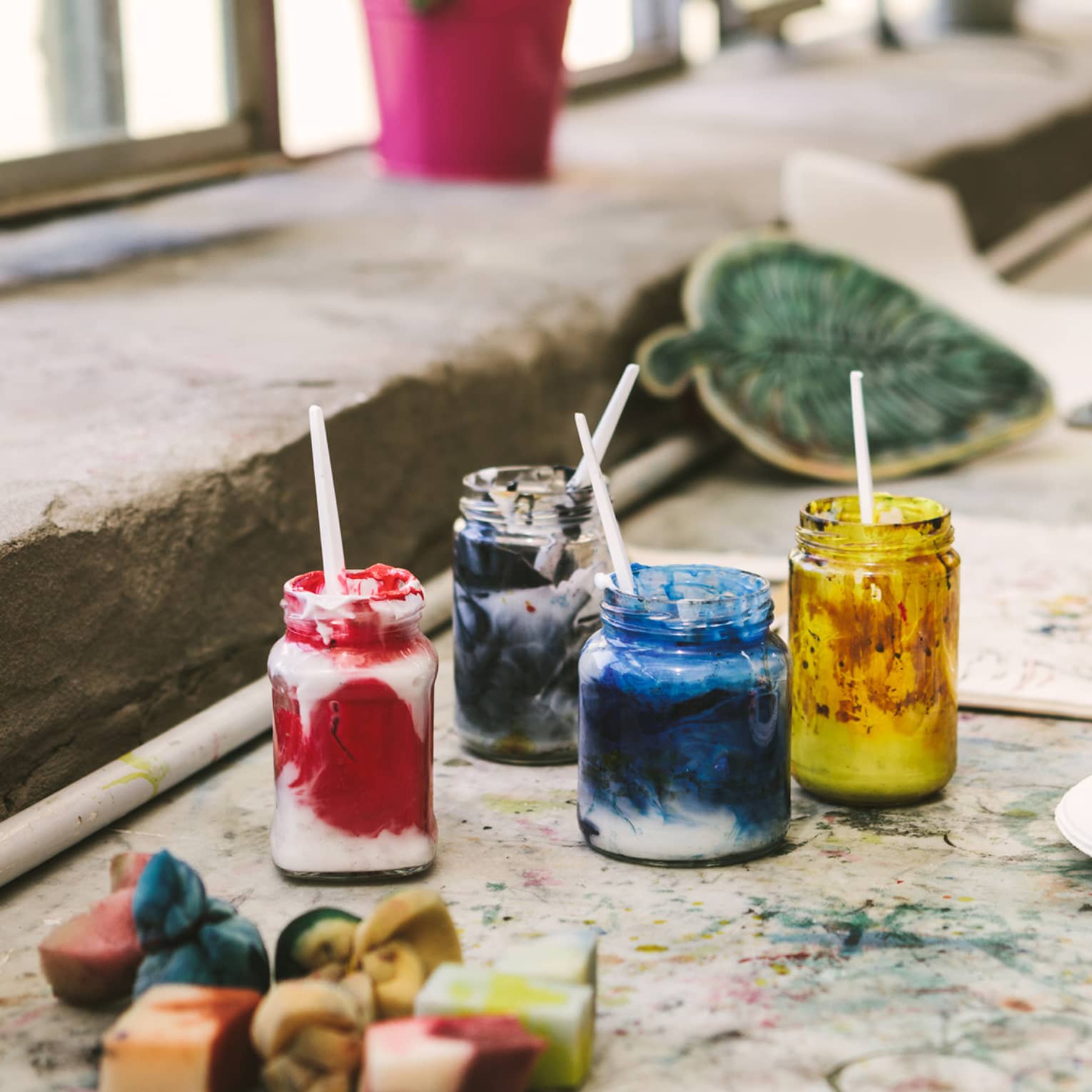 Close-up of a paint-splattered table covered in paper, jars of mixed paints and used painting sponges by a bright window.