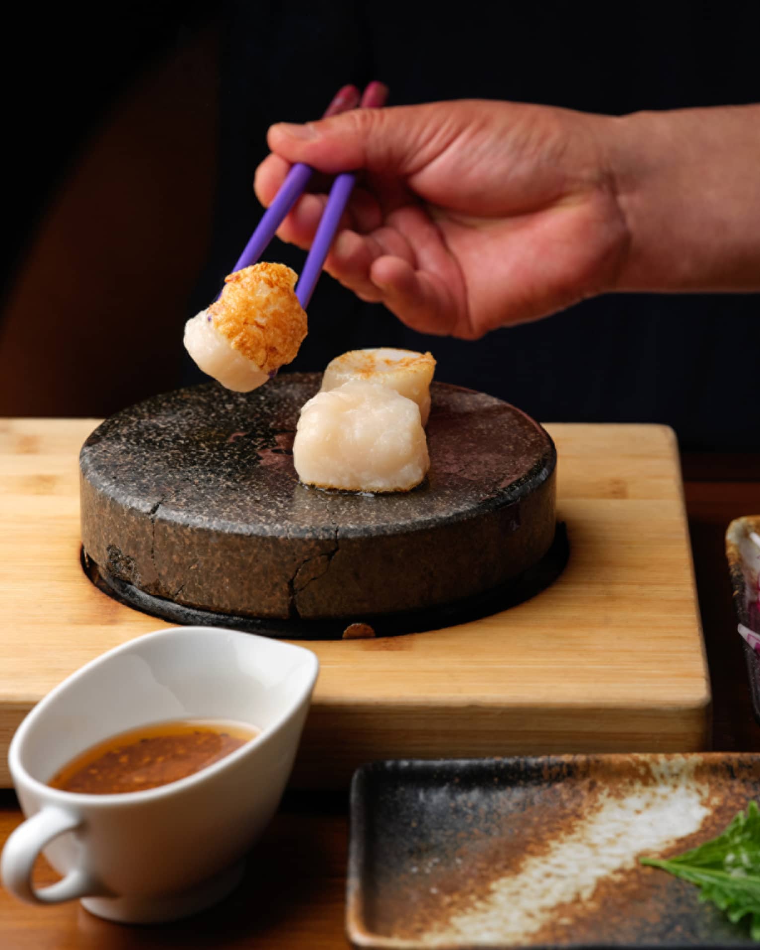 Close-up of a hand picking up a seared scallop with chopsticks from a stone tray, near small dishes of sauce and fresh sides.