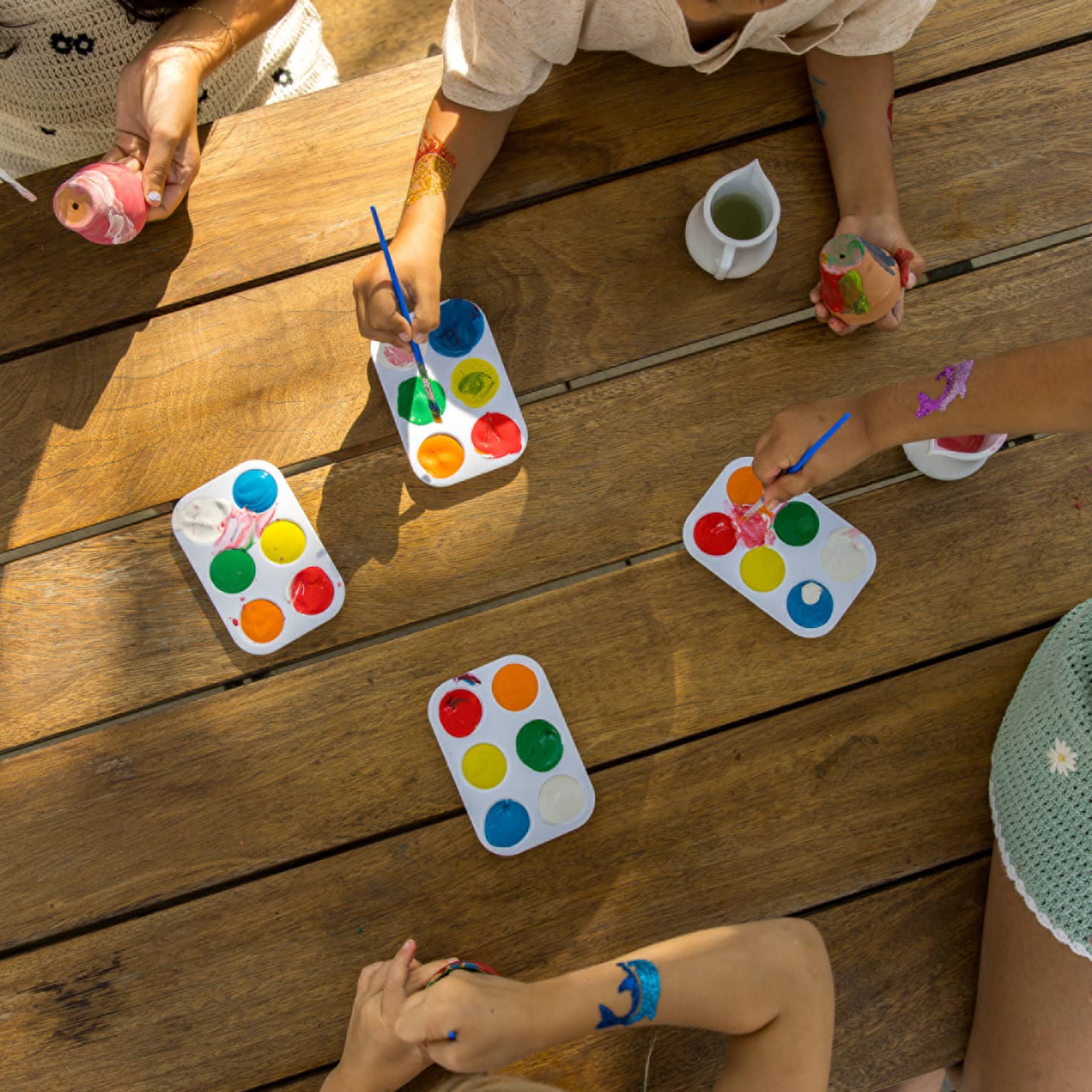 Overhead view of kids hands reaching into palettes of paint with their paint brushes on a wooden table