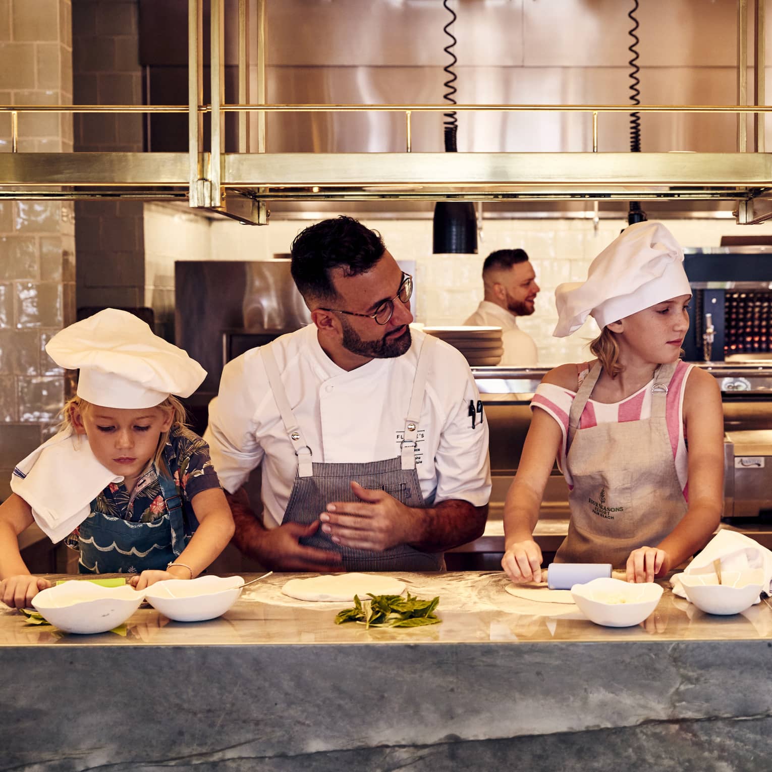 A chef standing at a counter teaching children wearing aprons and chef hats how to roll out pizza dough using rolling pins.