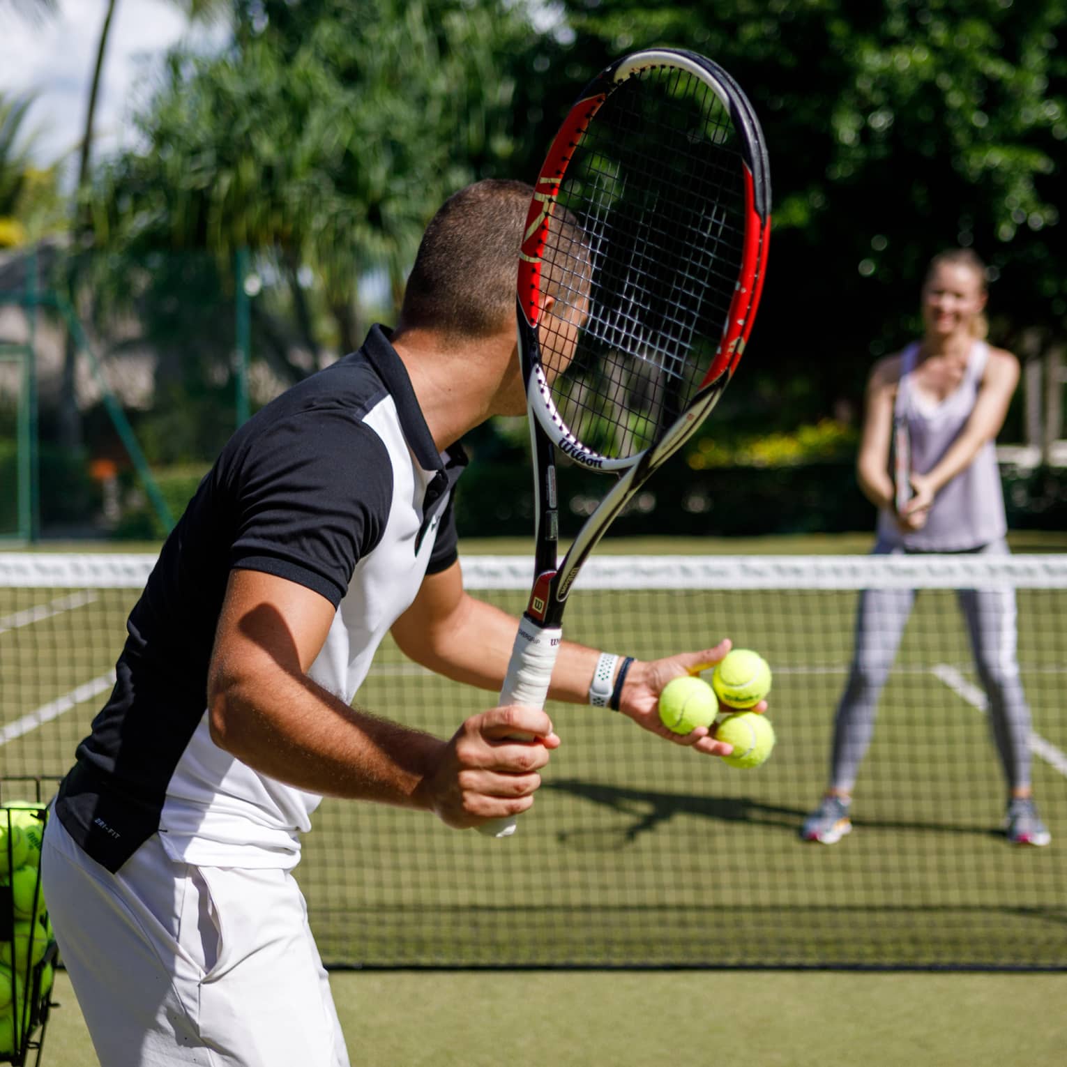 Couple playing tennis on court, man prepares to serve tennis balls to woman ready to swing